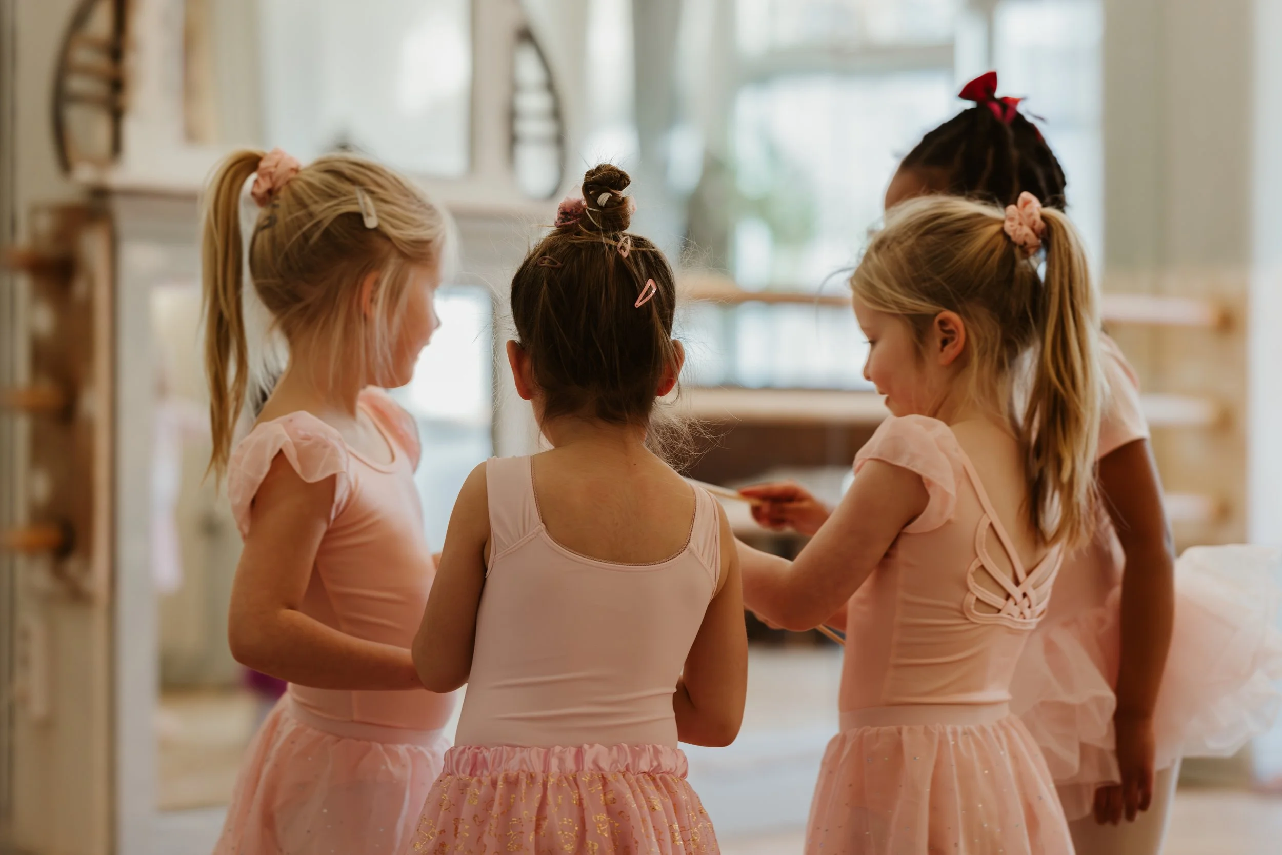 Vijf jonge meisjes in balletoutfits die samen in een dansstudio staan en naar elkaar kijken. Balletpakje, balletschool Rotterdam, kleuterdans, peuterdans, balletles, muziek, dans, Rotterdam west