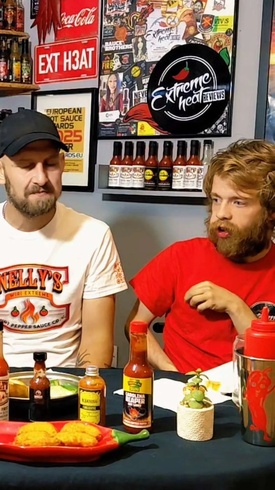 Two men sitting at a table with hot food and hot sauce bottles in front of them, decor with hot sauce signs, stickers, and posters on the wall behind them.