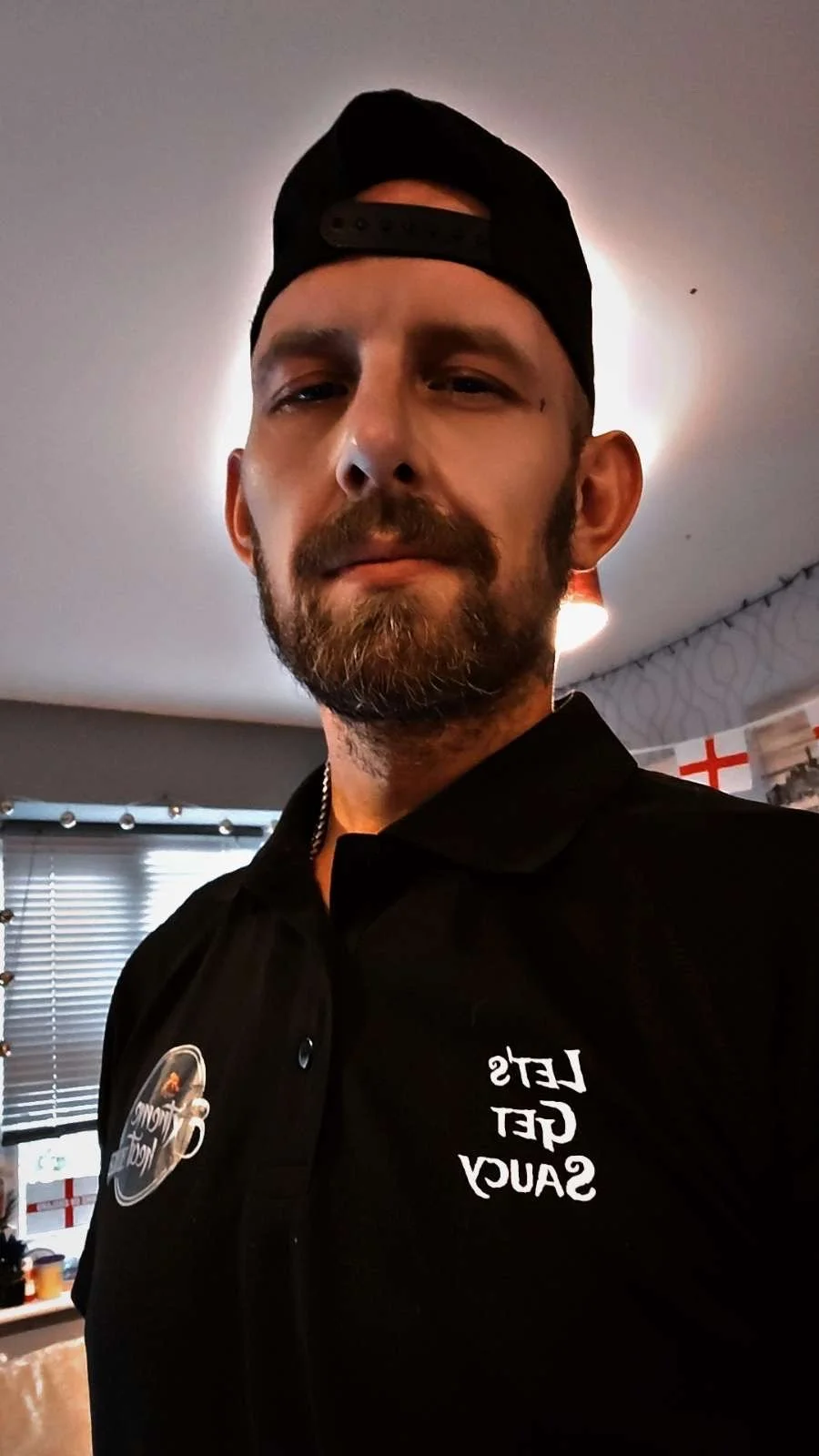 A man with a beard and mustache, wearing a black shirt with the text 'Let's Get Saucy' and a button, a black cap worn backward, in a room with blinds and a poster of the England flag in the background.