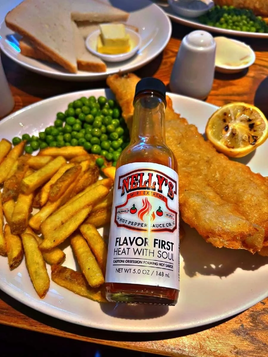 Fried fish fillet with lemon half, green peas, and French fries on a white plate, with a bottle of hot pepper sauce, bread with butter, and some condiments in the background.