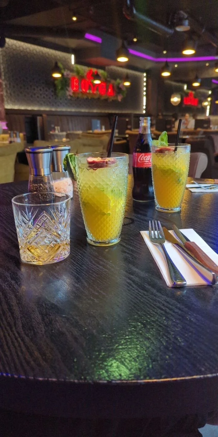 Two yellow cocktails with lime garnishes and two black straws on a dark wooden table in a restaurant. Also on the table are a glass with a drink, a set of salt and pepper shakers, a Coca-Cola bottle, and a fork and knife on a napkin. The background s