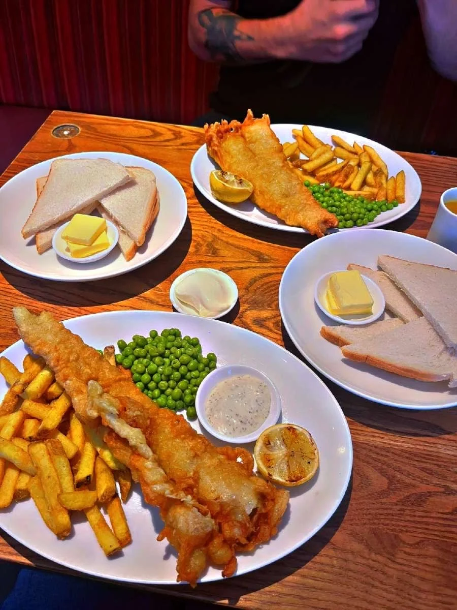 Two plates of fish and chips with green peas, tartar sauce, and grilled lemon halves, along with slices of white bread with butter on a wooden table in a restaurant setting.