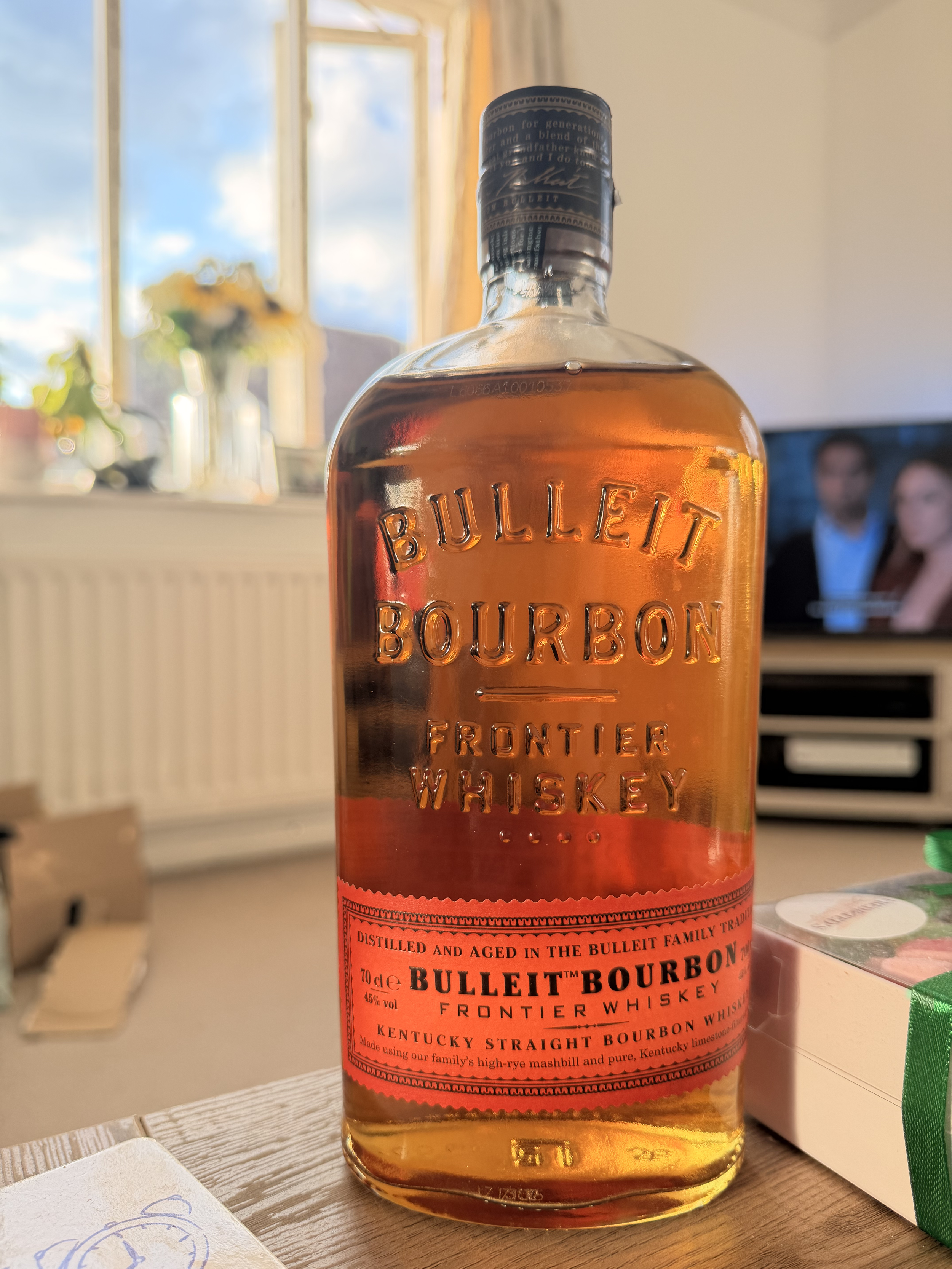 A bottle of Bulleit Bourbon frontler whiskey on a wooden table with a window, a TV showing two people, and other items in the background.