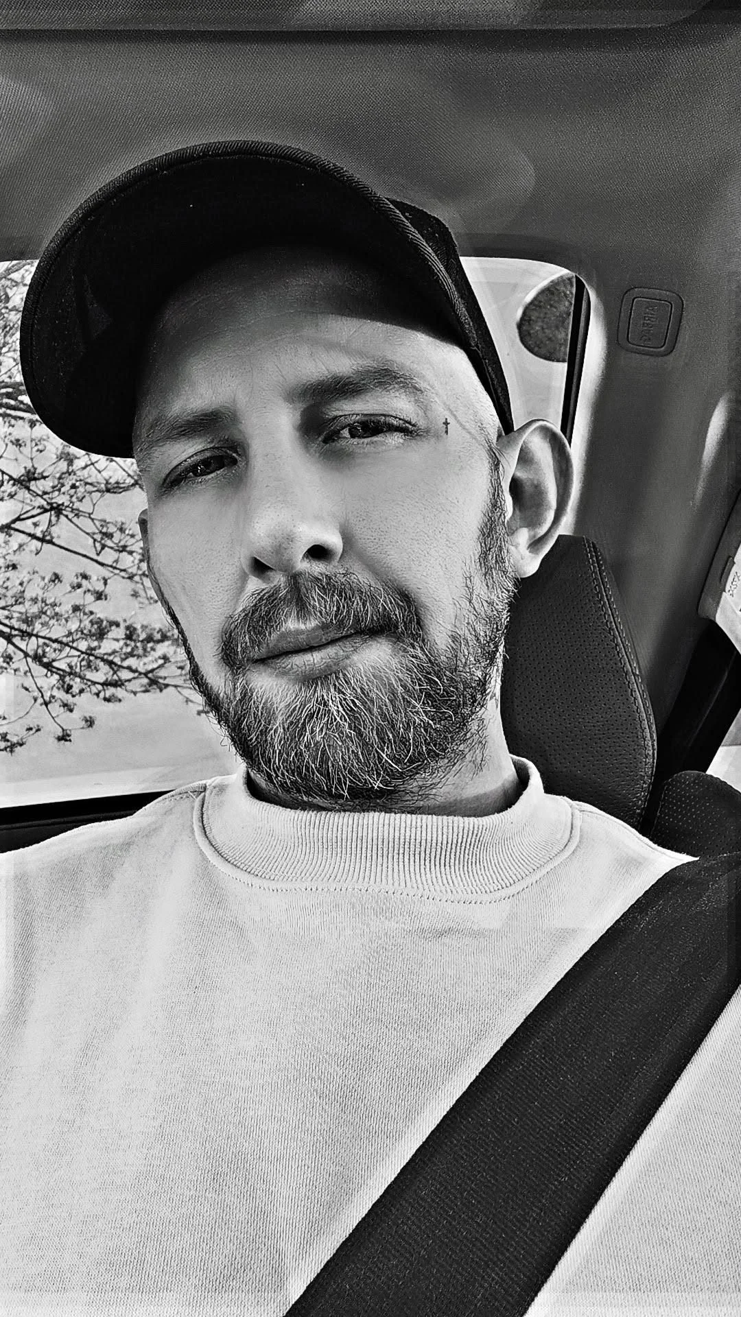 A man with a beard and mustache wearing a baseball cap and a light-colored sweater, sitting in a car, taking a selfie.