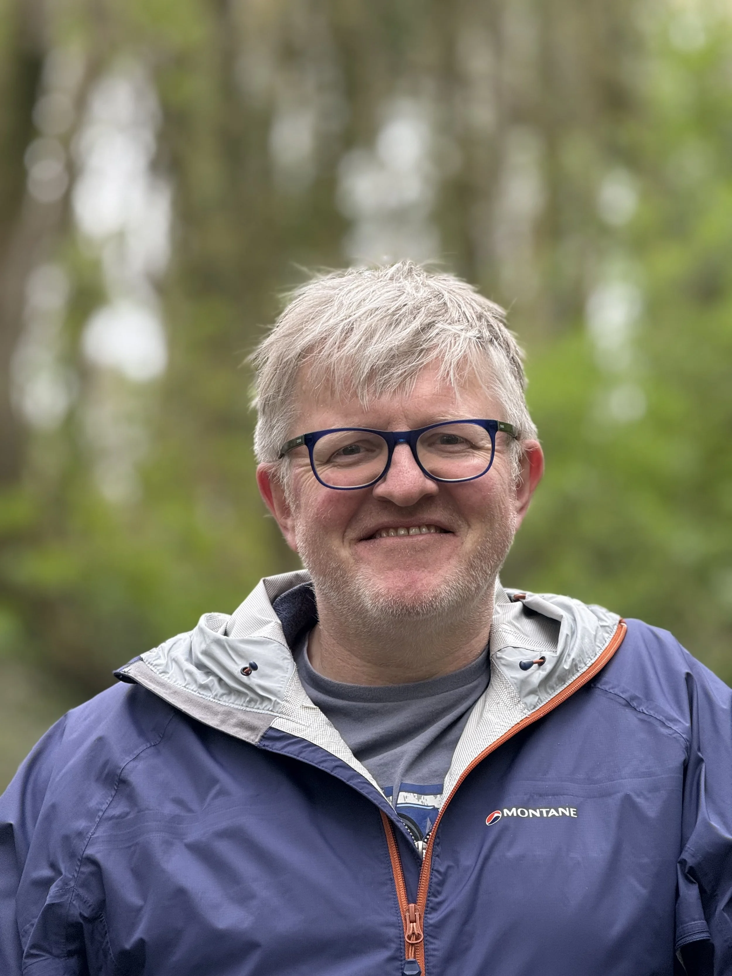 A smiling middle-aged man with gray hair, wearing glasses, a gray shirt, and a blue Montane jacket, standing outdoors in a forested area with blurred green trees in the background.