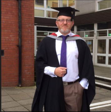A man in academic graduation attire, including a black cap and gown with a purple tie, standing outside a building with large windows and brick walls.