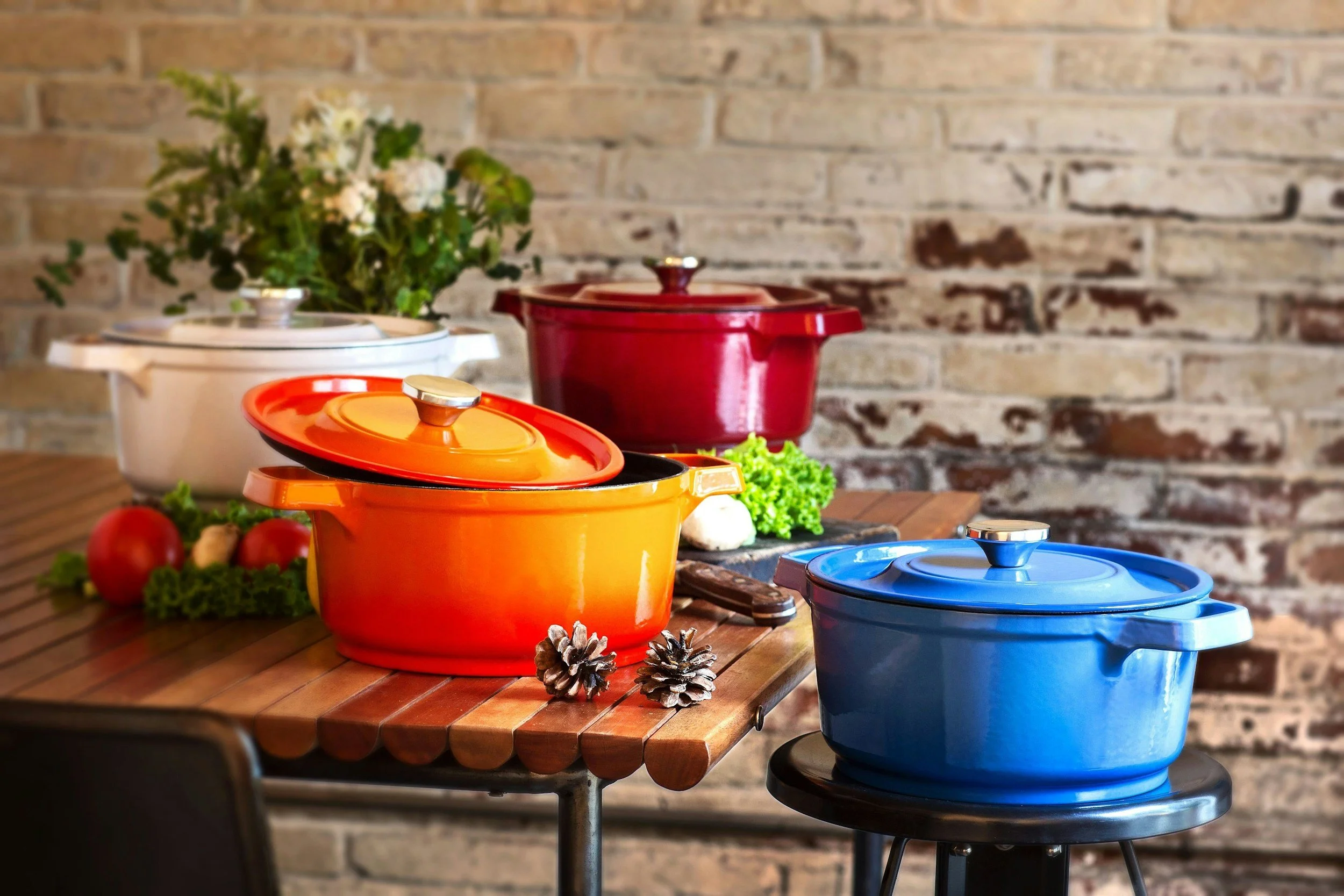 Colorful cast iron pots with lids on a wooden table, decorated with pinecones, tomatoes, and fresh greens, against a rustic brick wall background.