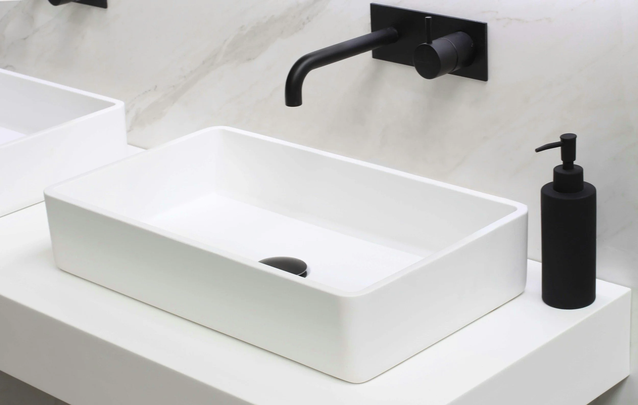 A modern bathroom sink setup with a rectangular white vessel sink, a black wall-mounted faucet, a black soap dispenser, and a light-colored marble or stone wall in the background.