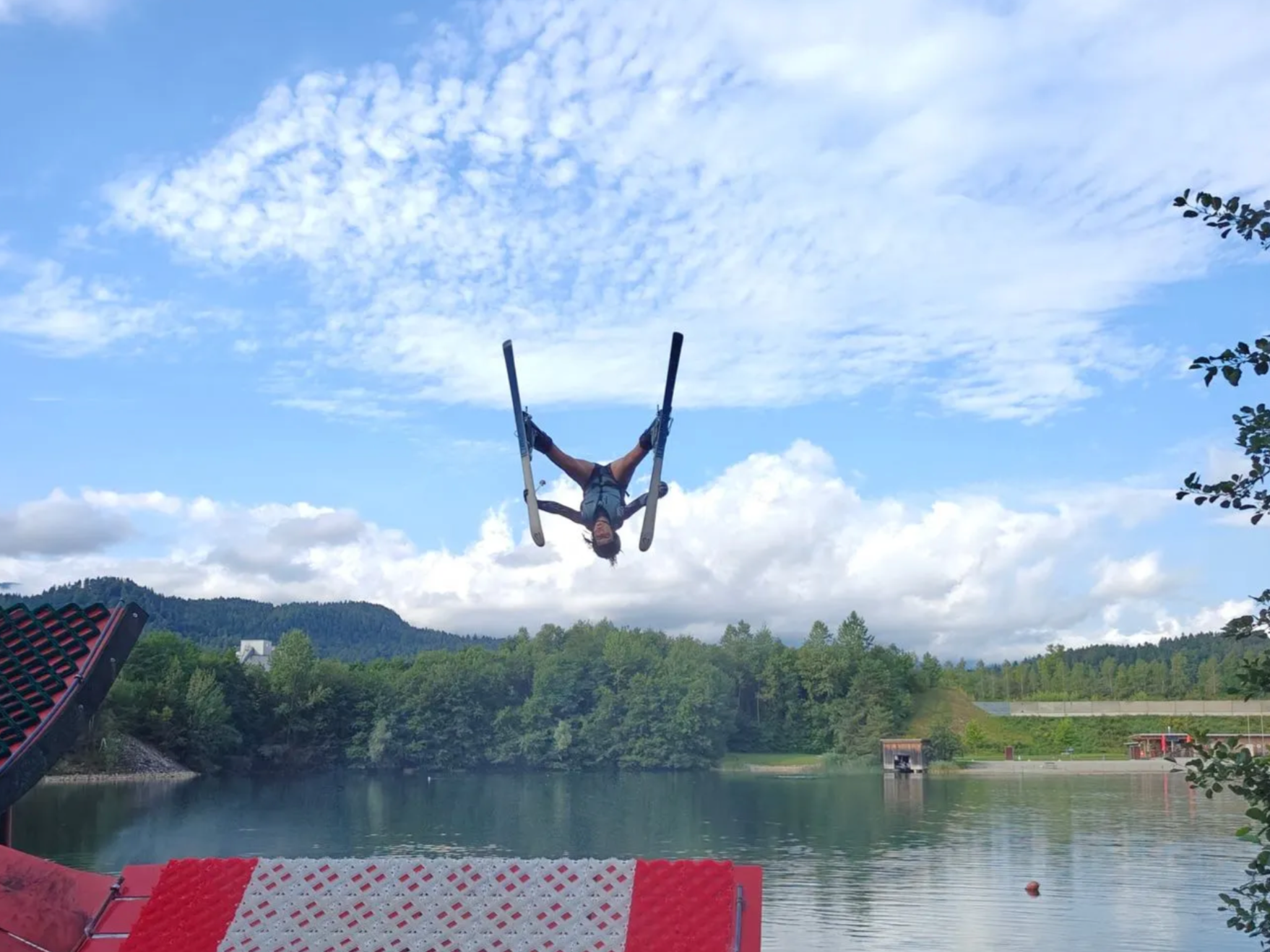 Person mid-air on a wakeboard upside down over a lake with a scenic mountain and cloudy sky background.