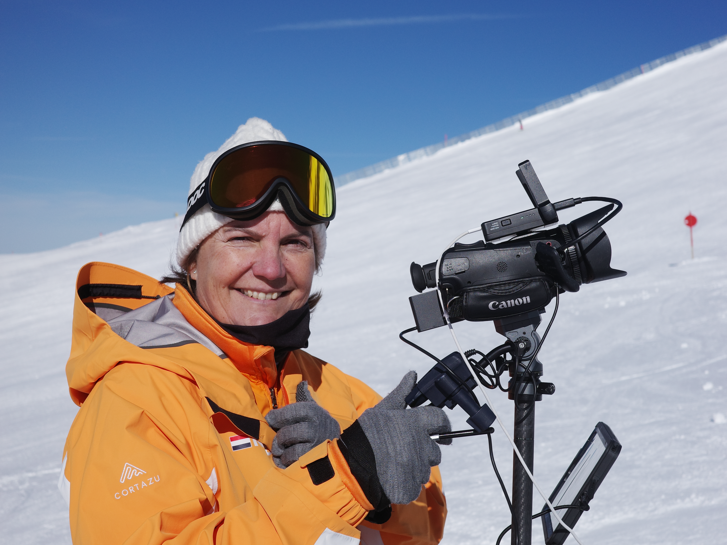 A woman in an orange jacket and white beanie with ski goggles is smiling while operating a video camera on a snowy mountain under a clear blue sky.