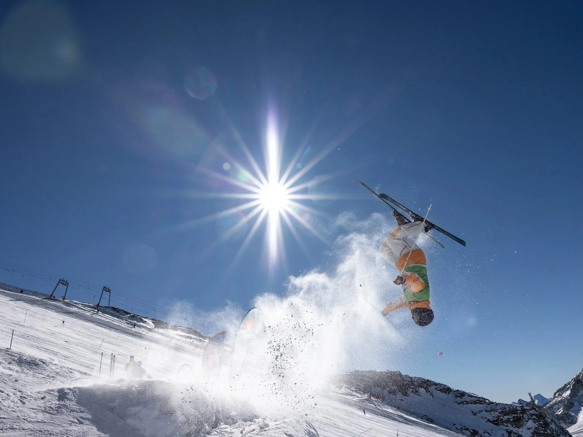 A skier performing a flip in mid-air on a snowy mountain under a clear blue sky with the sun shining brightly.