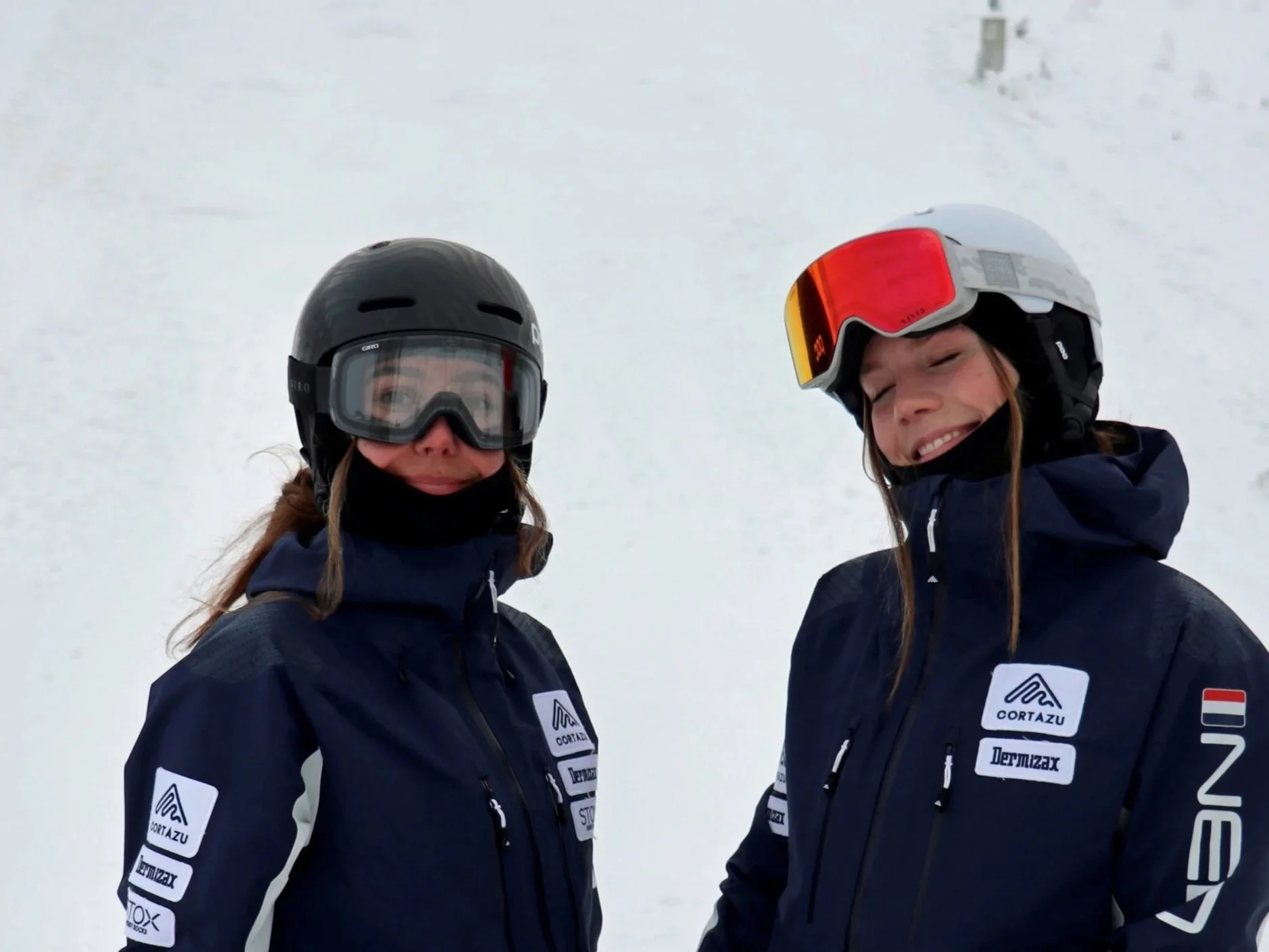 Two women wearing black helmets and winter gear standing on a snow-covered mountain. Both are smiling and wearing jackets with patches from Cortazu, Dermlux, and others.