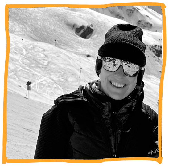 A woman wearing a beanie, sunglasses, and winter jacket, smiling on a snowy mountain.