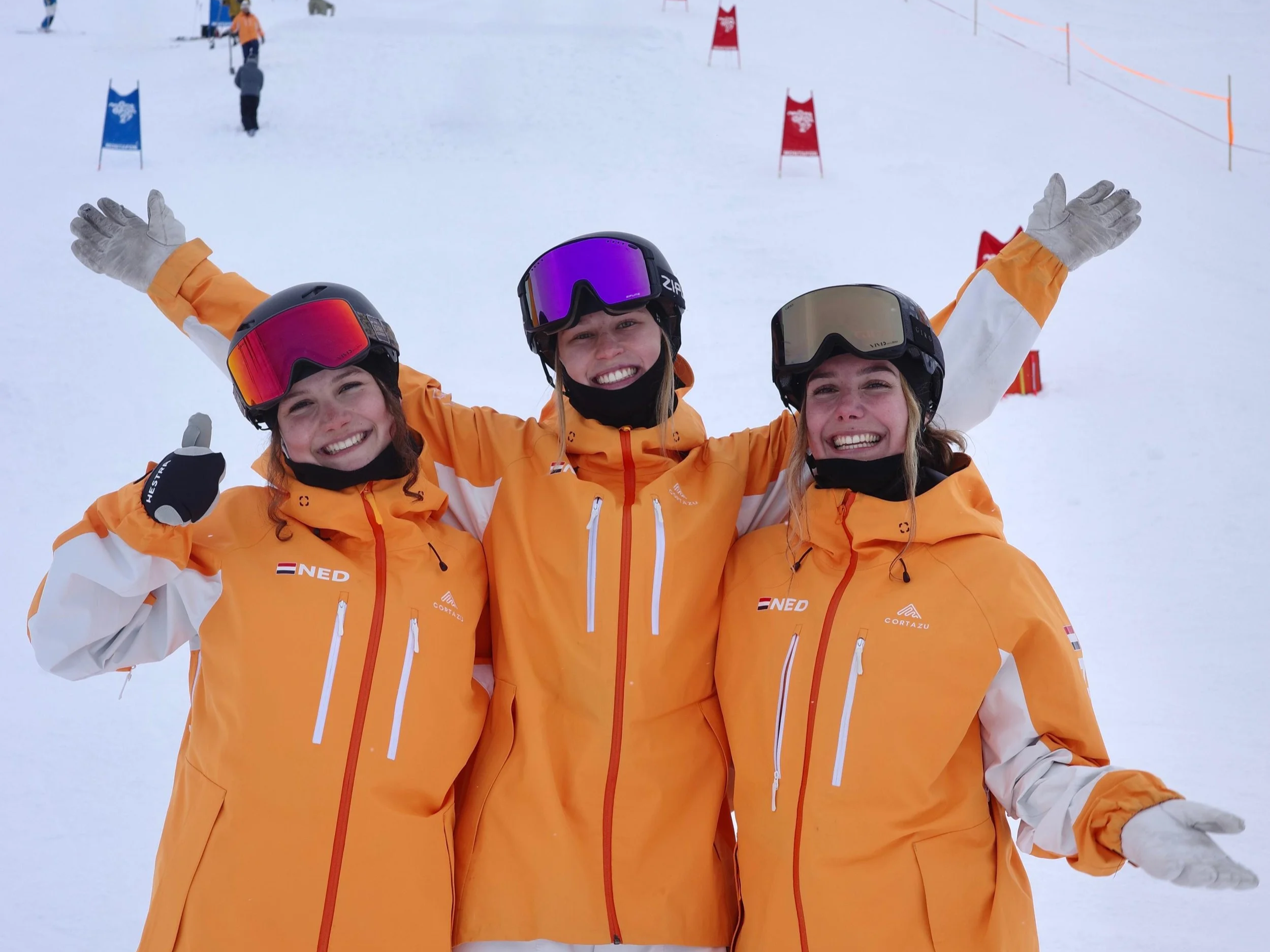 Three young female skiers in orange jackets and ski helmets smiling and celebrating on a snowy ski slope with flags and other skiers in the background.