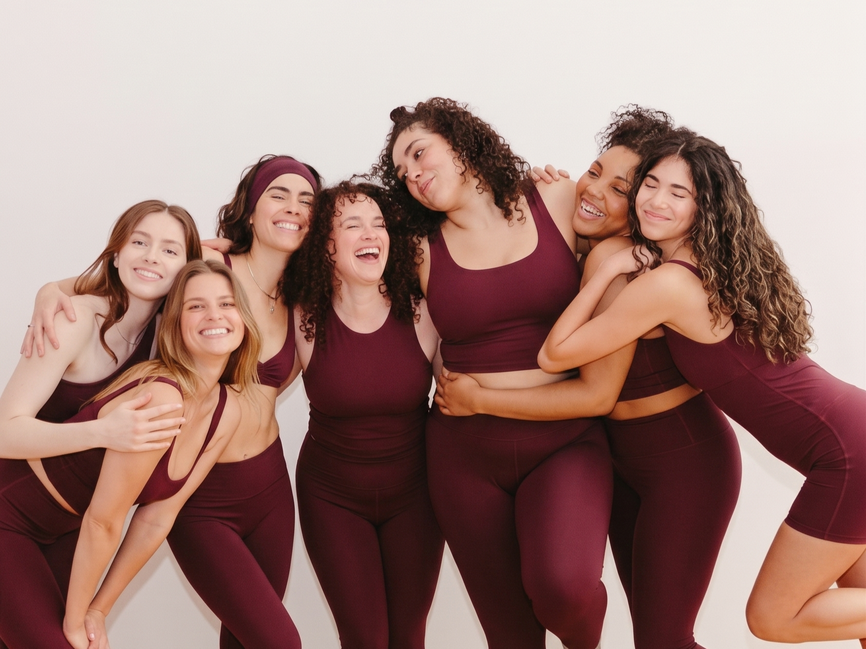 Groupe de sept femmes de différentes origines ethniques portant des vêtements de sport rouge bordeaux, en train de rire dans une ambiance chaleureuse