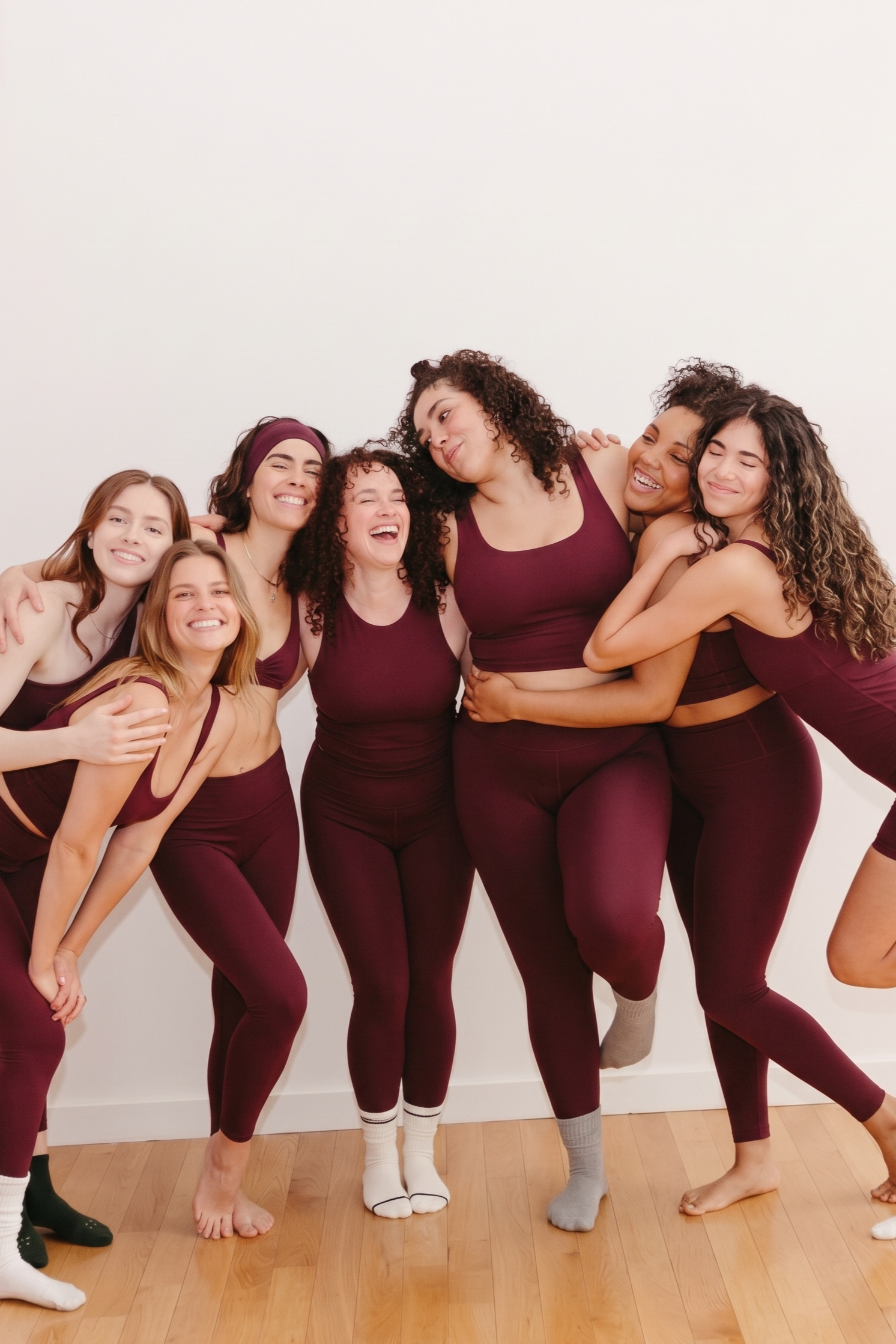 Groupe de sept femmes de différentes origines ethniques portant des vêtements de sport rouge bordeaux, en train de rire dans une ambiance chaleureuse