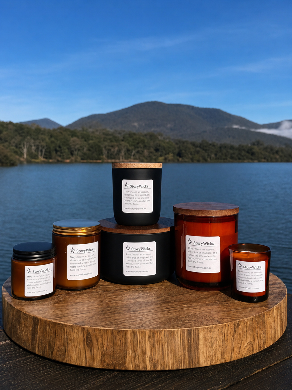 Six candles of various sizes with white labels on a wooden round platform, overlooking a lake with green trees and mountains in the background.