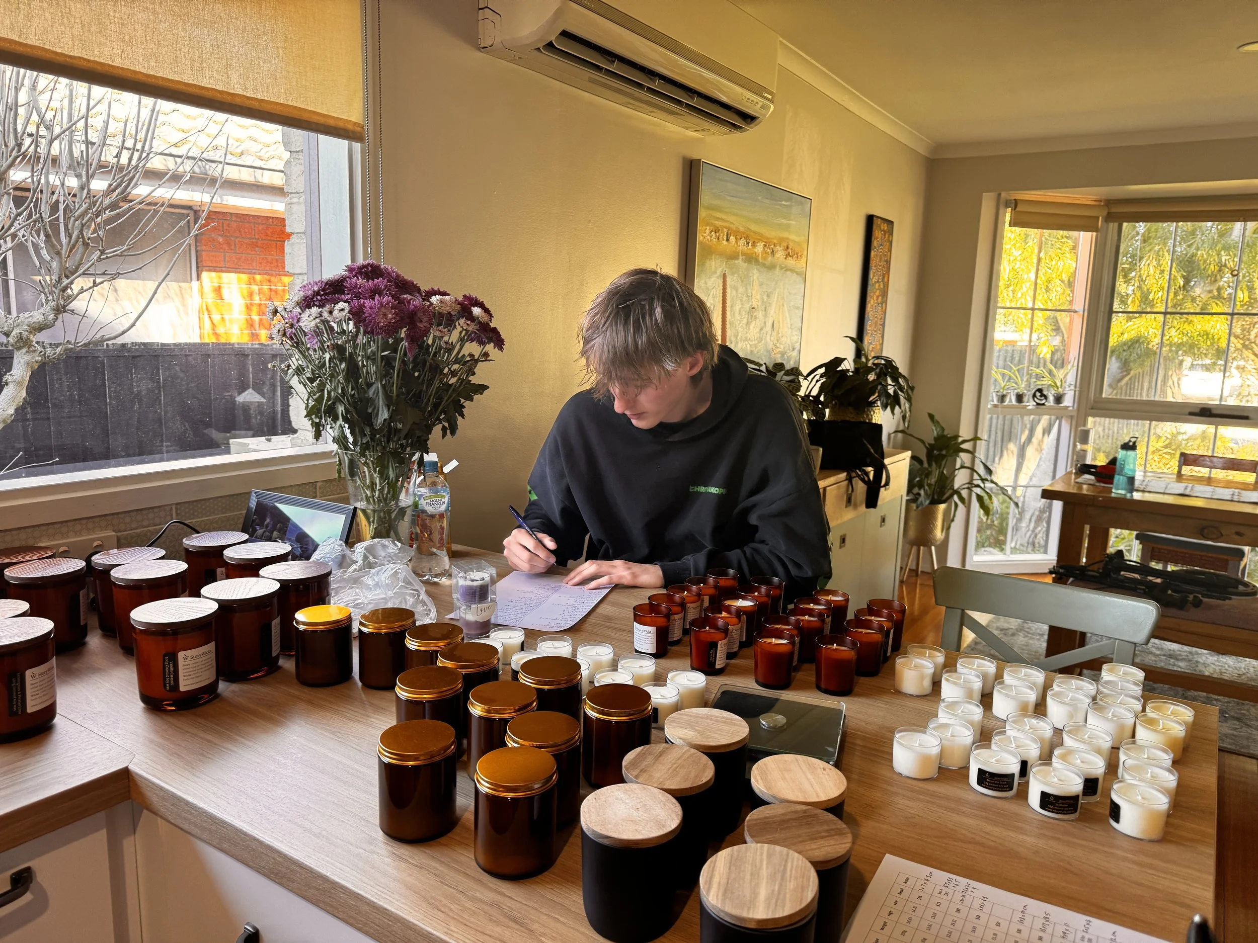 A person with gray hair wearing a black hoodie sitting at a table with numerous candles in brown and white containers, some with wooden lids, and lined up on the table. There is a large window with blinds, a bouquet of purple flowers, and framed art on the yellow wall in the background.