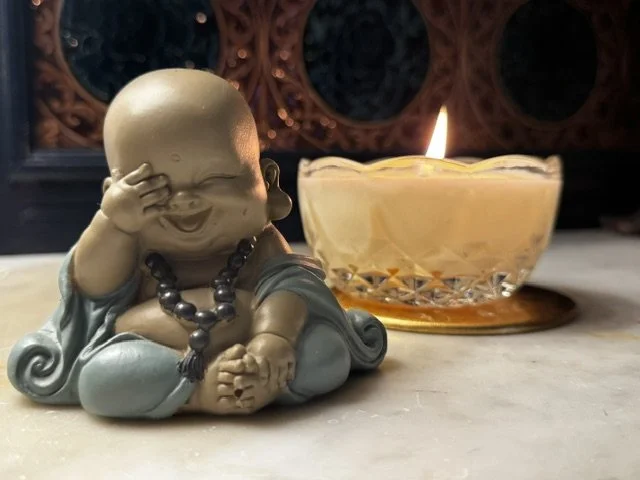 A small laughing Buddha statue wearing a beaded necklace, sitting cross-legged with one hand covering its eye and the other hand resting on its knee, placed next to a lit candle in a glass holder.