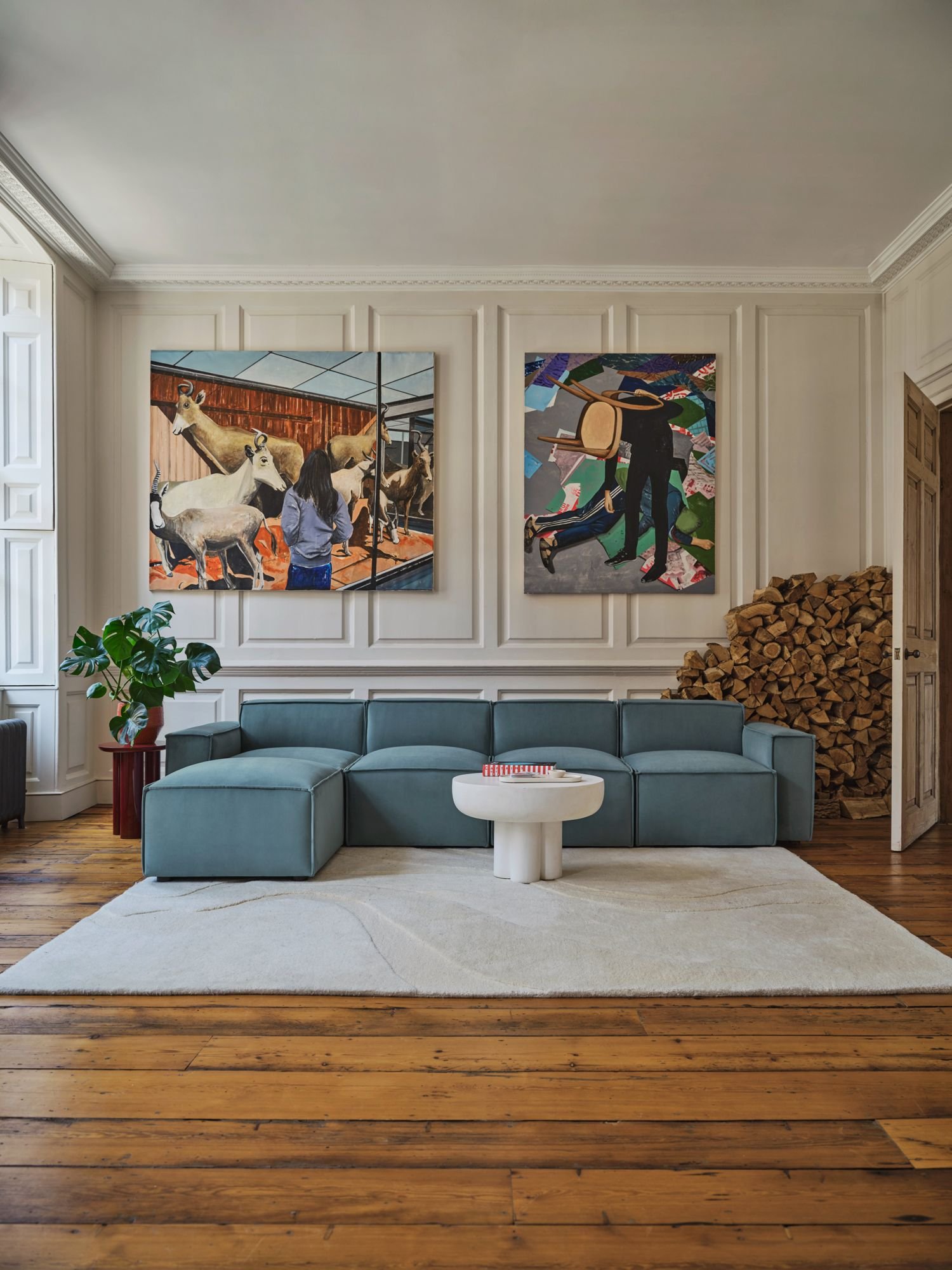 Living room with a teal sofa, white coffee table, green potted plant, wood floor, white walls with paneling, two large paintings on the wall, and a stack of firewood in the corner.