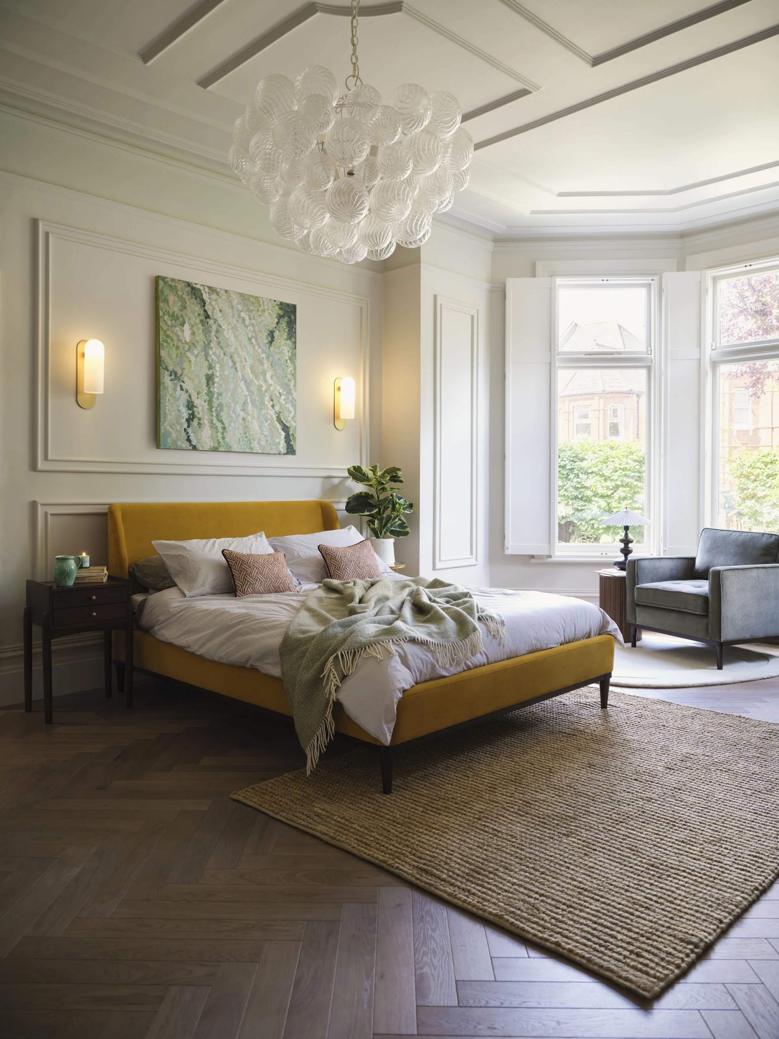 A bedroom with a yellow upholstered bed, nightstand with books and decorative items, wall art with abstract green and white pattern, large window with white frame, gray armchair, hanging glass chandelier, and a woven rug on dark wood flooring.