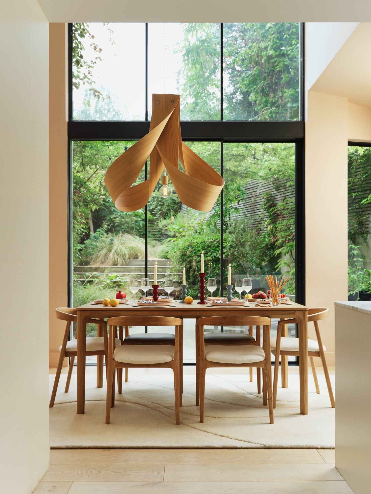Modern dining room with a wooden table set for a meal, surrounded by chairs, inside a room with large floor-to-ceiling windows showing greenery outside. Hanging from the ceiling is a contemporary wooden chandelier, and the table is decorated with candles, apples, glasses, and dinnerware.