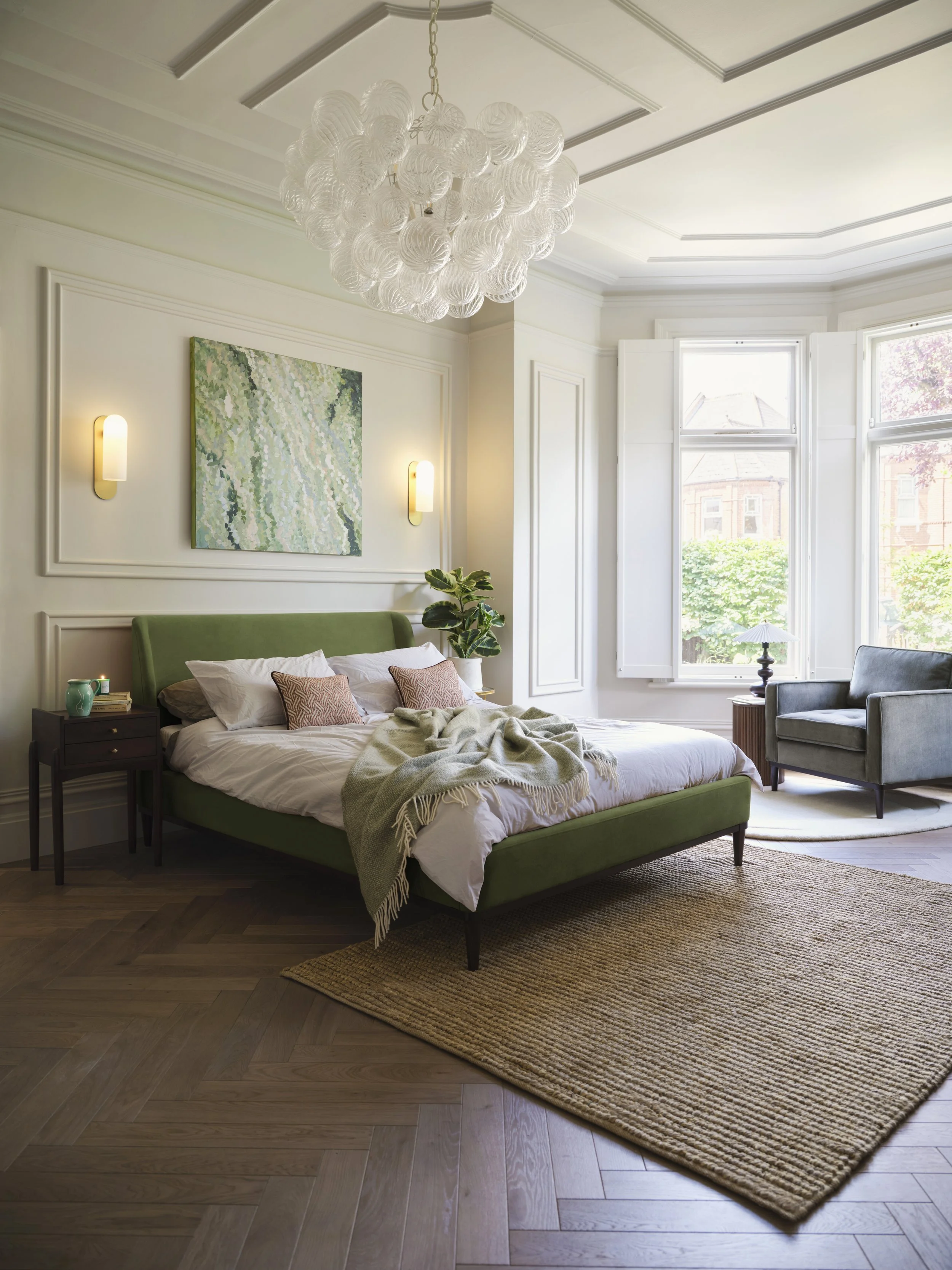 A bedroom featuring a bed with white sheets, a green headboard, and decorative pillows. Beside the bed is a dark wood nightstand with a teal vase, books, and candles. The room has two large windows with white trim, allowing natural light. There's a gray armchair, a small side table with a lamp, a potted plant, a unique white chandelier hanging from the ceiling, and a brown woven rug on the wooden floor. An abstract painting is on the wall above the bed, illuminated by two wall sconces.