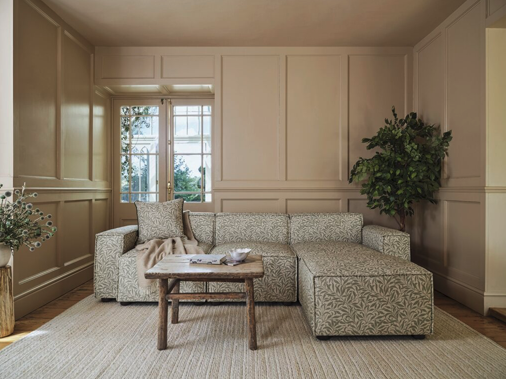 Living room with a cream-colored, patterned sectional sofa, a wooden coffee table, a potted plant, and large windows allowing natural light, with beige walls and wood flooring.