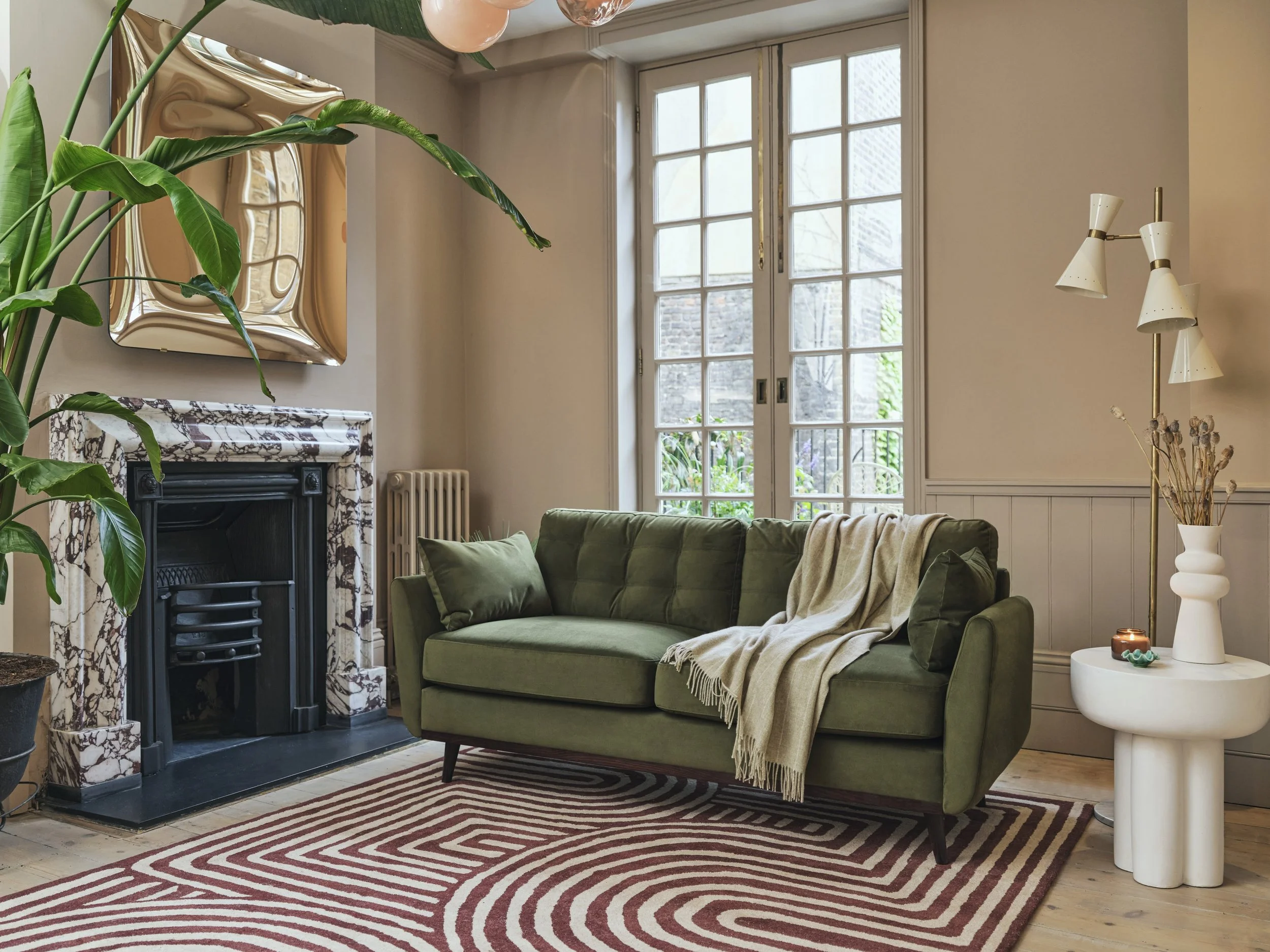 A cozy living room with a green velvet sofa, a striped red and white rug, a white side table with decorative objects, and a large window with grid panes allowing natural light. A plant with large green leaves is near a fireplace with a marble surround, and a modern floor lamp is on the right side of the image.
