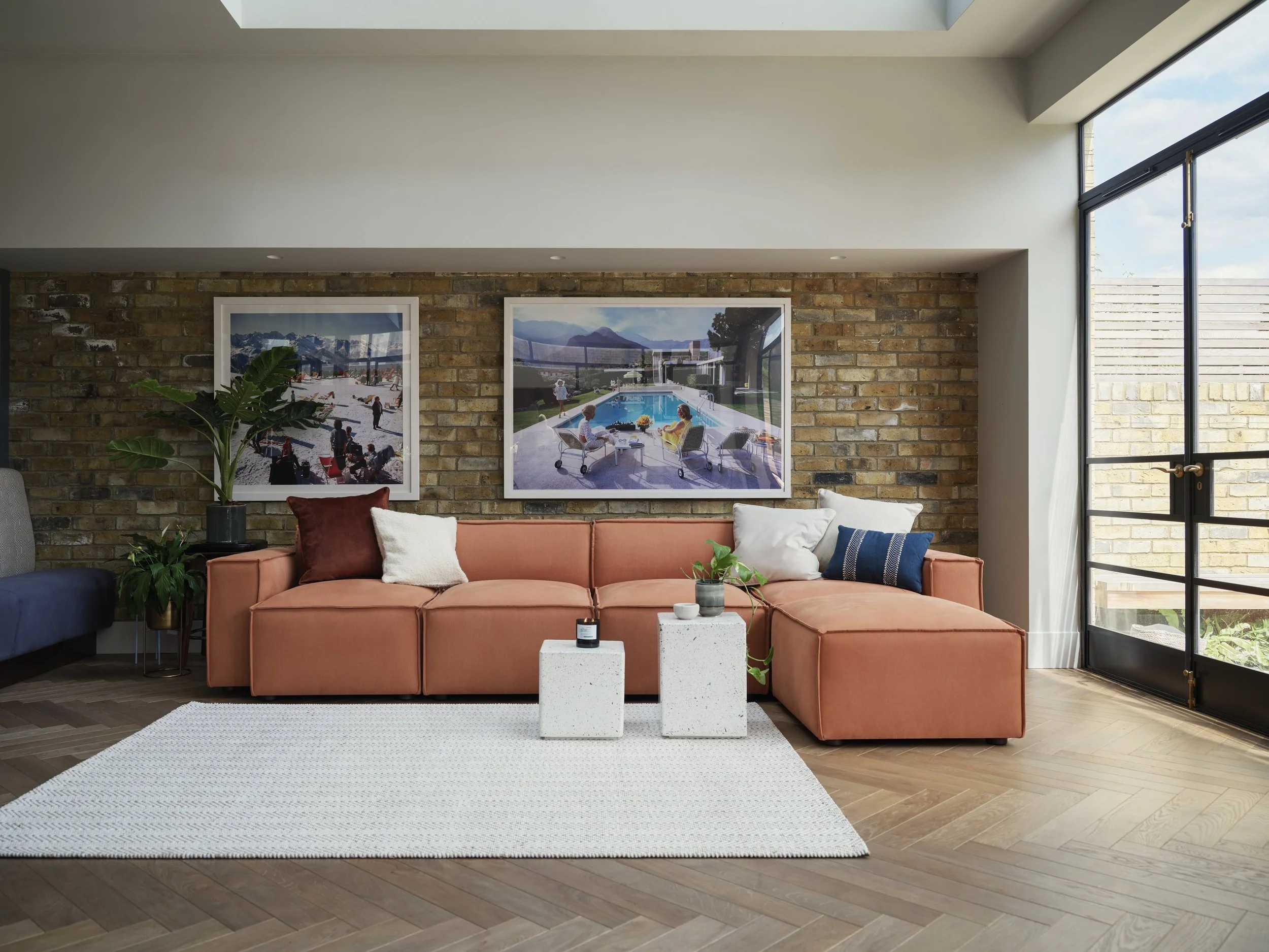 Living room with brick accent wall, large window, pink sofa, artwork of outdoor scenes, green plants, white and black tables, white area rug, wood flooring.