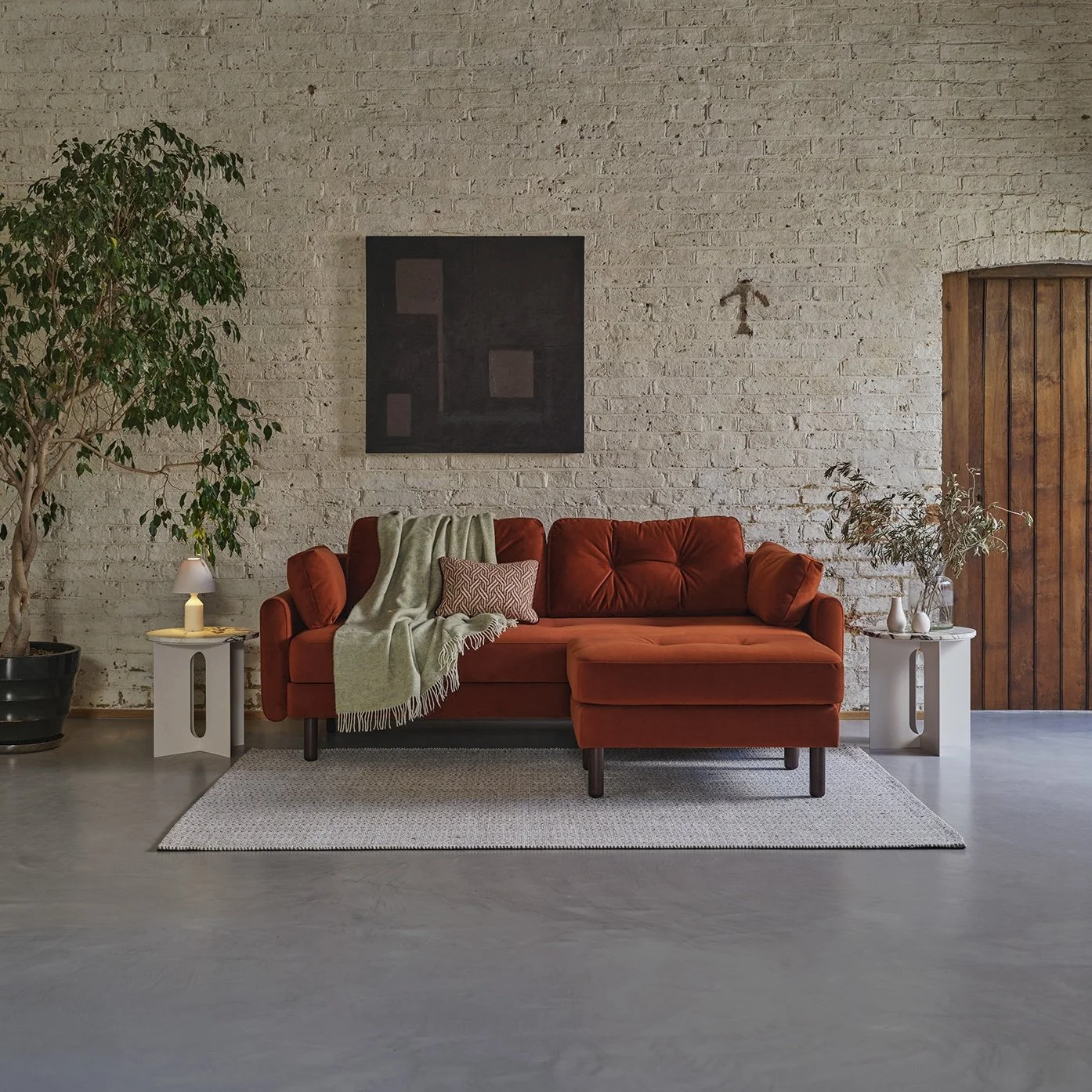 Living room with a brick wall, orange sofa, two white side tables, a large green plant, a small lamp, and white vases. A painting hangs on the wall, and a throw blanket and pillow are on the sofa.