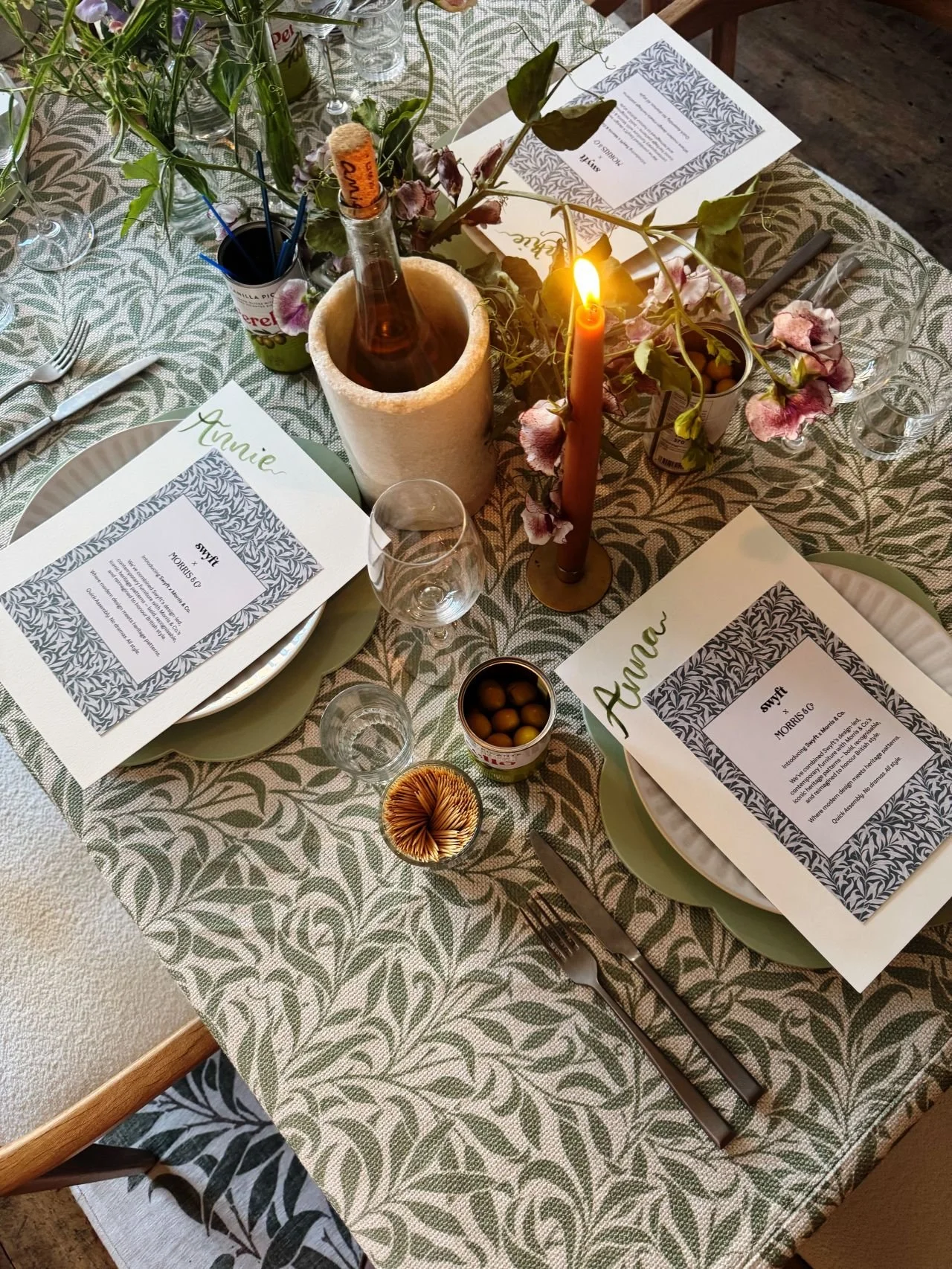 A decorated dining table set with papers, glasses, plates, and cutlery, featuring a lit candle, a bottle in a ice holder, and floral arrangements with pink flowers and green leaves.