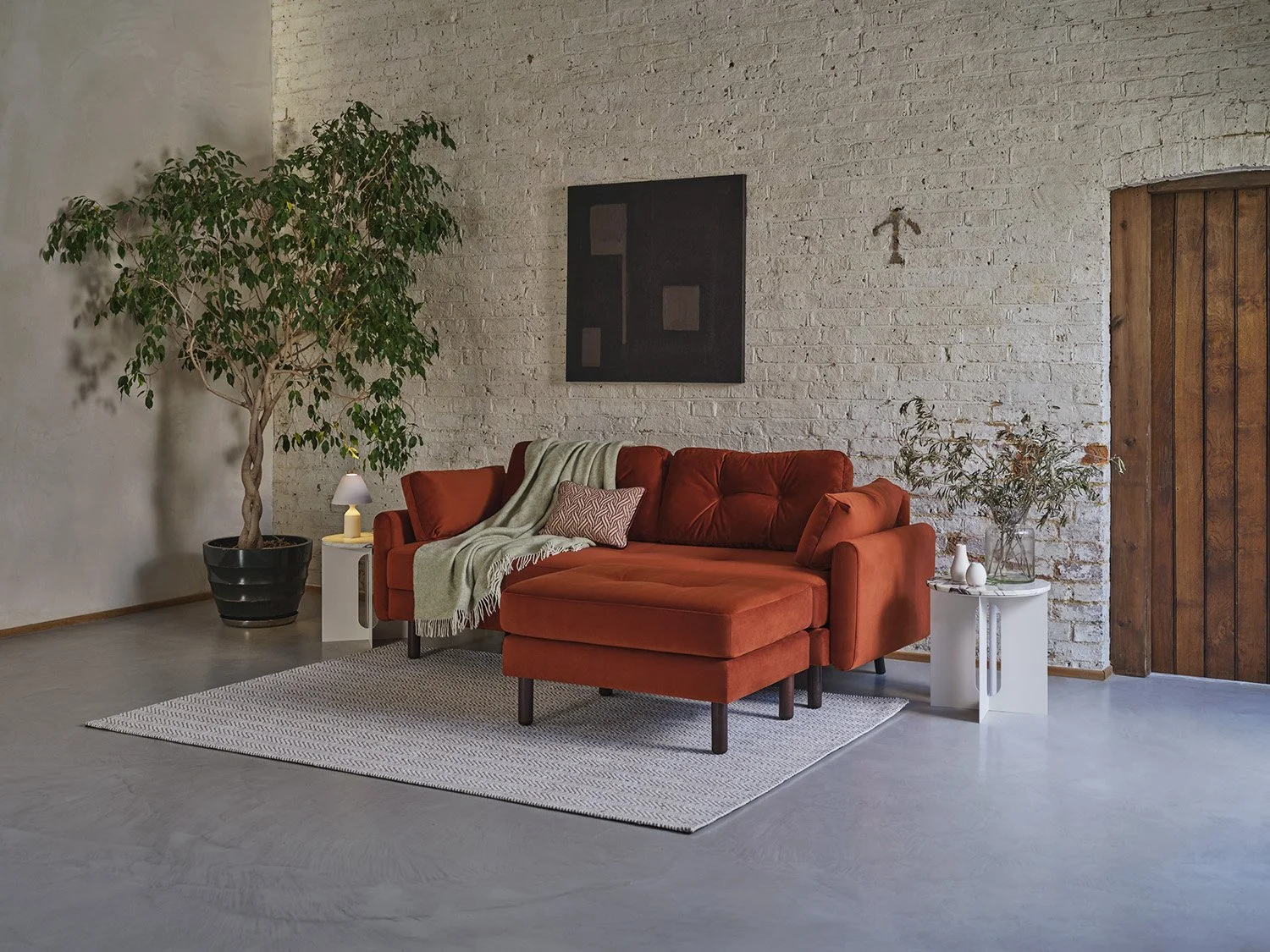 Living room with a large red sectional sofa, a small side table with a white lamp, a large potted tree, and a white side table with vases and dried plants, against a white brick wall and wooden door.