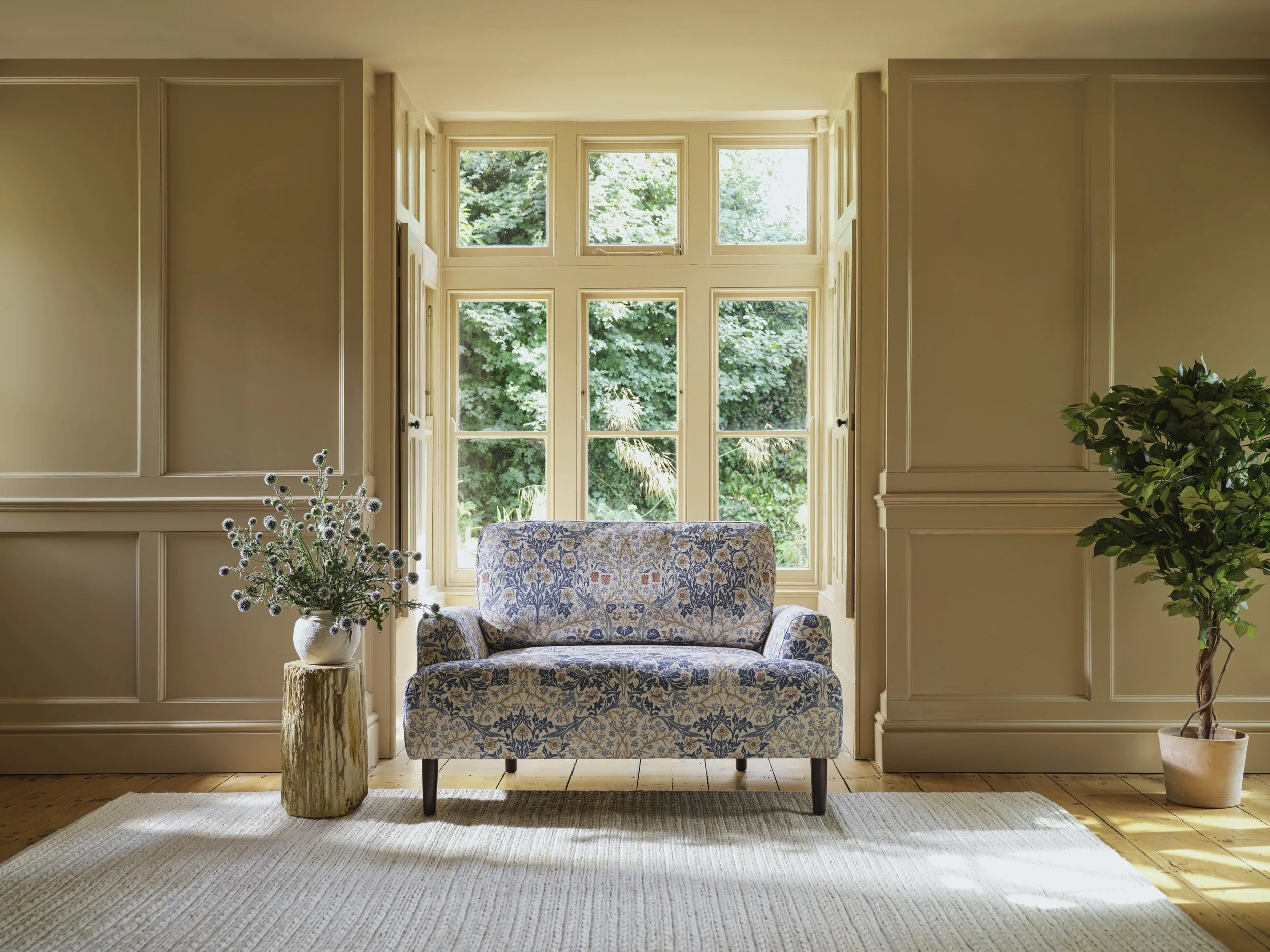 A cozy living room with beige walls and a large window, decorated with floral-patterned furniture, a potted plant, and a vase of flowers, with sunlight streaming through the window.