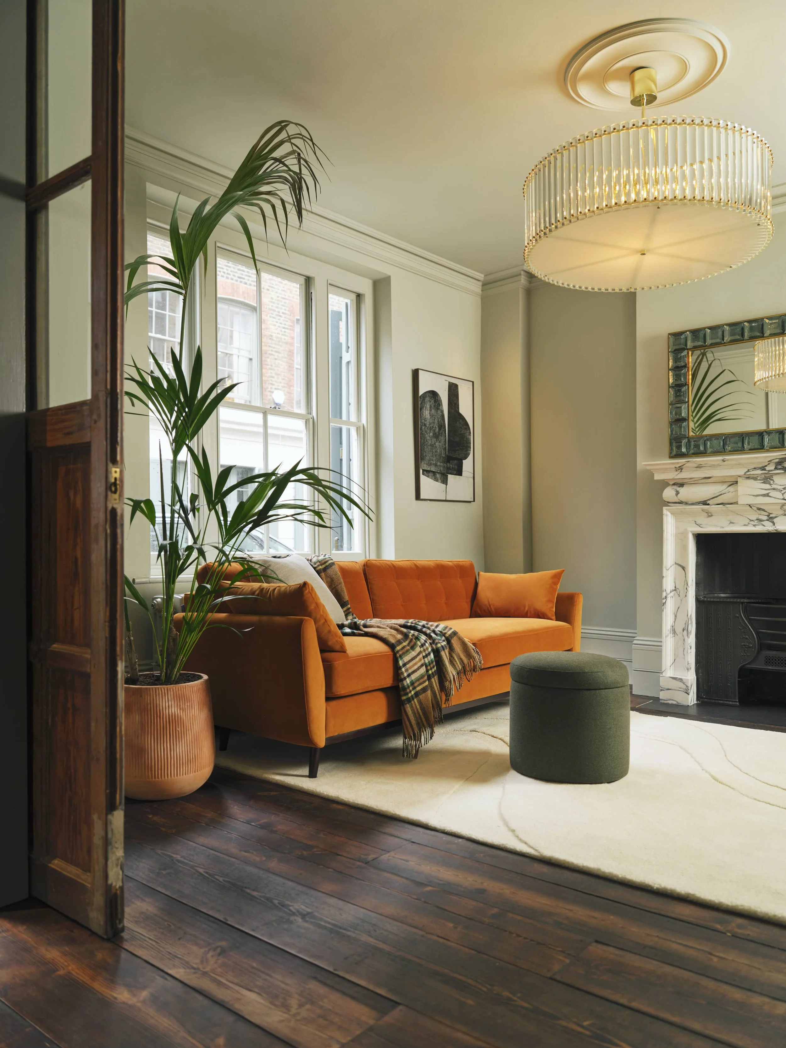A cozy living room with a bright orange sofa, a green round ottoman, tall green potted plant, black and white abstract art on the wall, large window with natural light, marble fireplace, and a modern chandelier overhead.