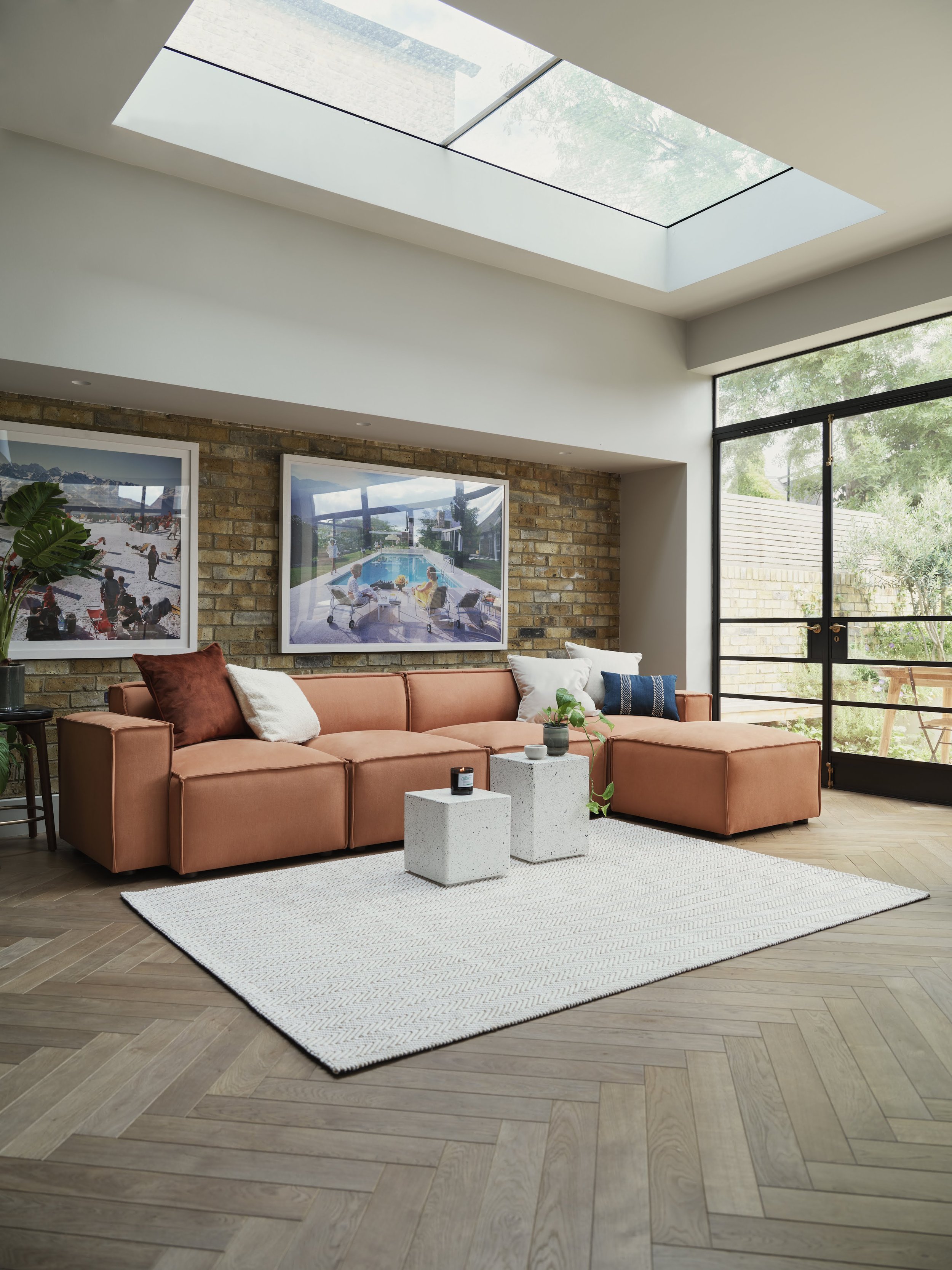 Living room with a pink sectional sofa, white and rust-colored pillows, two concrete side tables, a white textured rug, a brick accent wall with framed pictures, a potted plant, large sliding glass door, and a high, rectangular skylight in the ceiling allowing natural light.