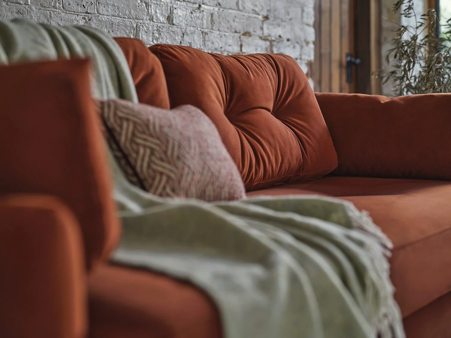 Close-up of a red velvet sofa with cushions and a throw blanket, against a white brick wall and wooden door.