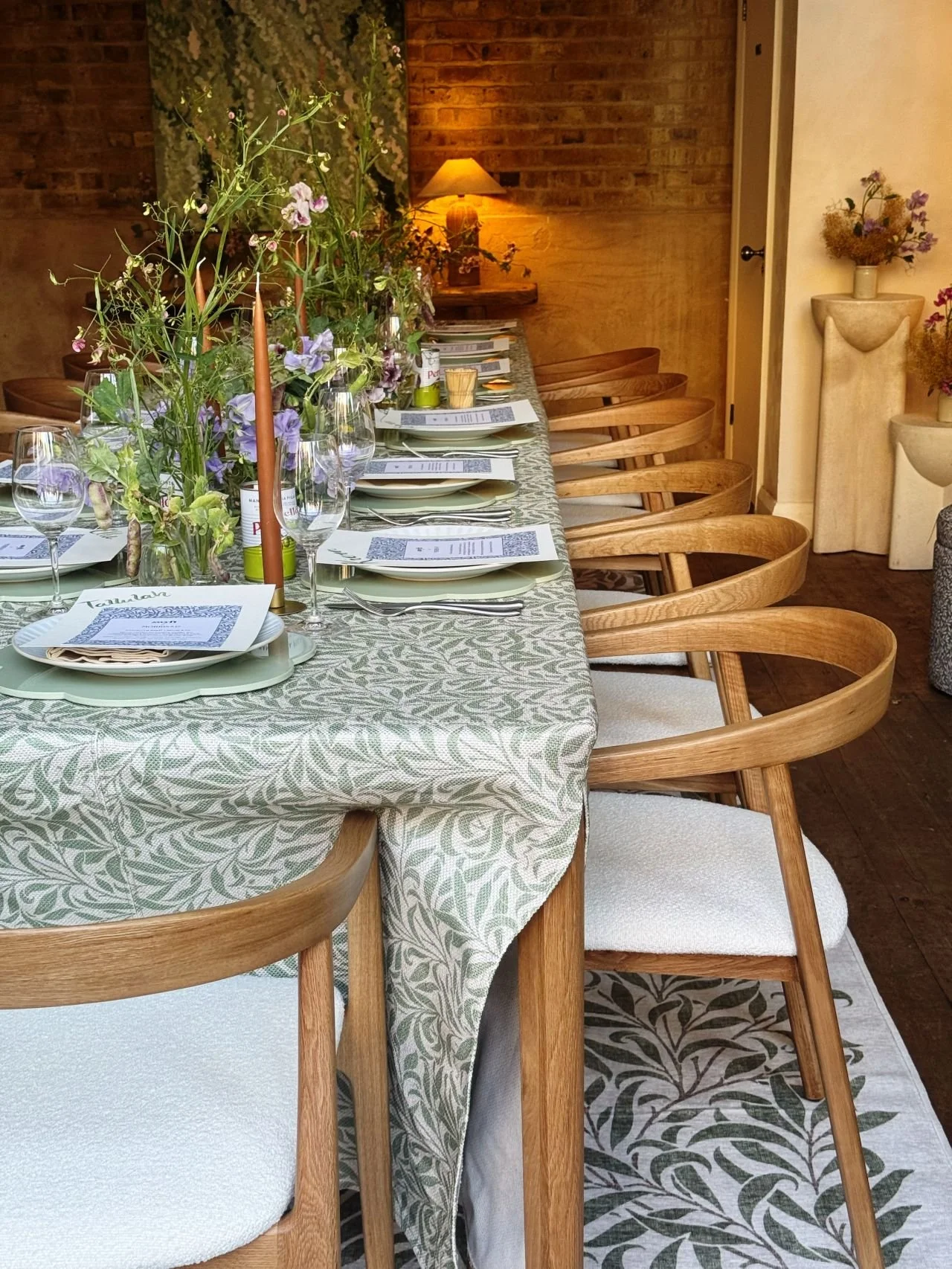 A long dining table with a light green patterned tablecloth, set with plates, napkins, silverware, wine glasses, and colorful flower centerpieces. Wooden chairs with cream cushions surround the table. The background includes brick and wood panel walls, a small lamp, and decorative vases with flowers.