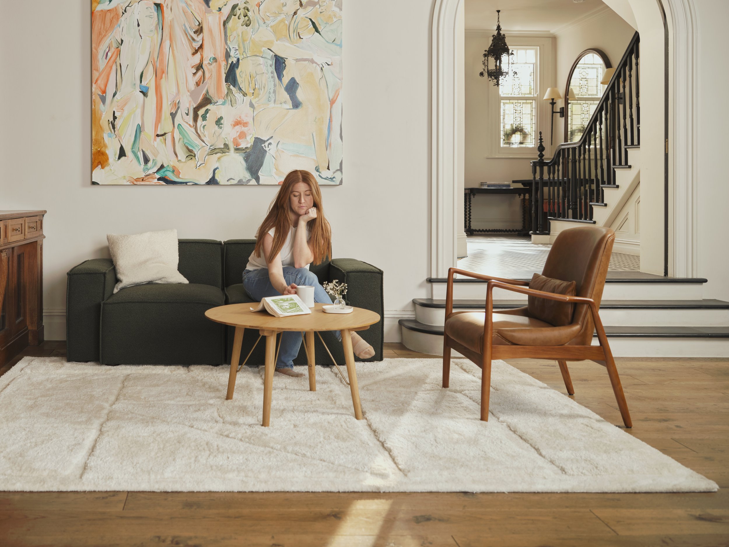 A woman with long red hair sitting on a dark green couch in a living room, holding a white mug, with an open book on a small round wooden coffee table in front of her. The room has a cream-colored carpet, a cream pillow on the couch, and a large abstract painting with pastel colors on the wall behind her. There is a staircase with black railings and two windows with stained glass design in the background.
