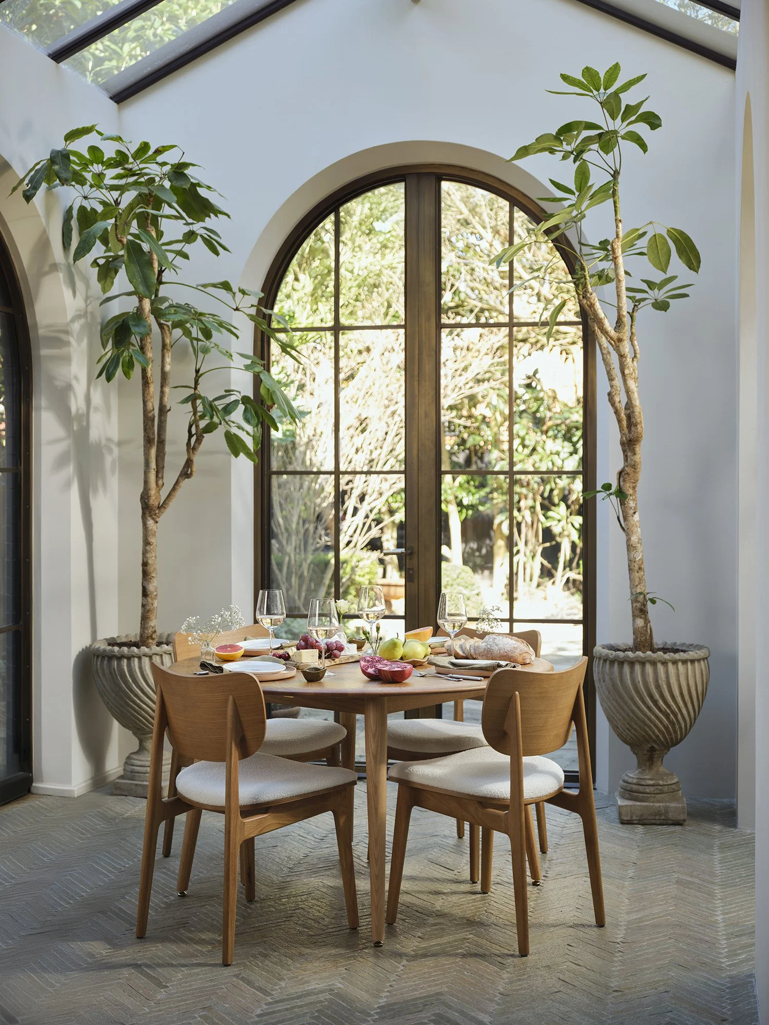 A dining area with a round wooden table set with plates, glasses, and fruits. Four wooden chairs with white cushions surround the table. Two large potted trees in decorative urns flank the area. A large arched window and glass ceiling let in natural light, revealing a garden outside.