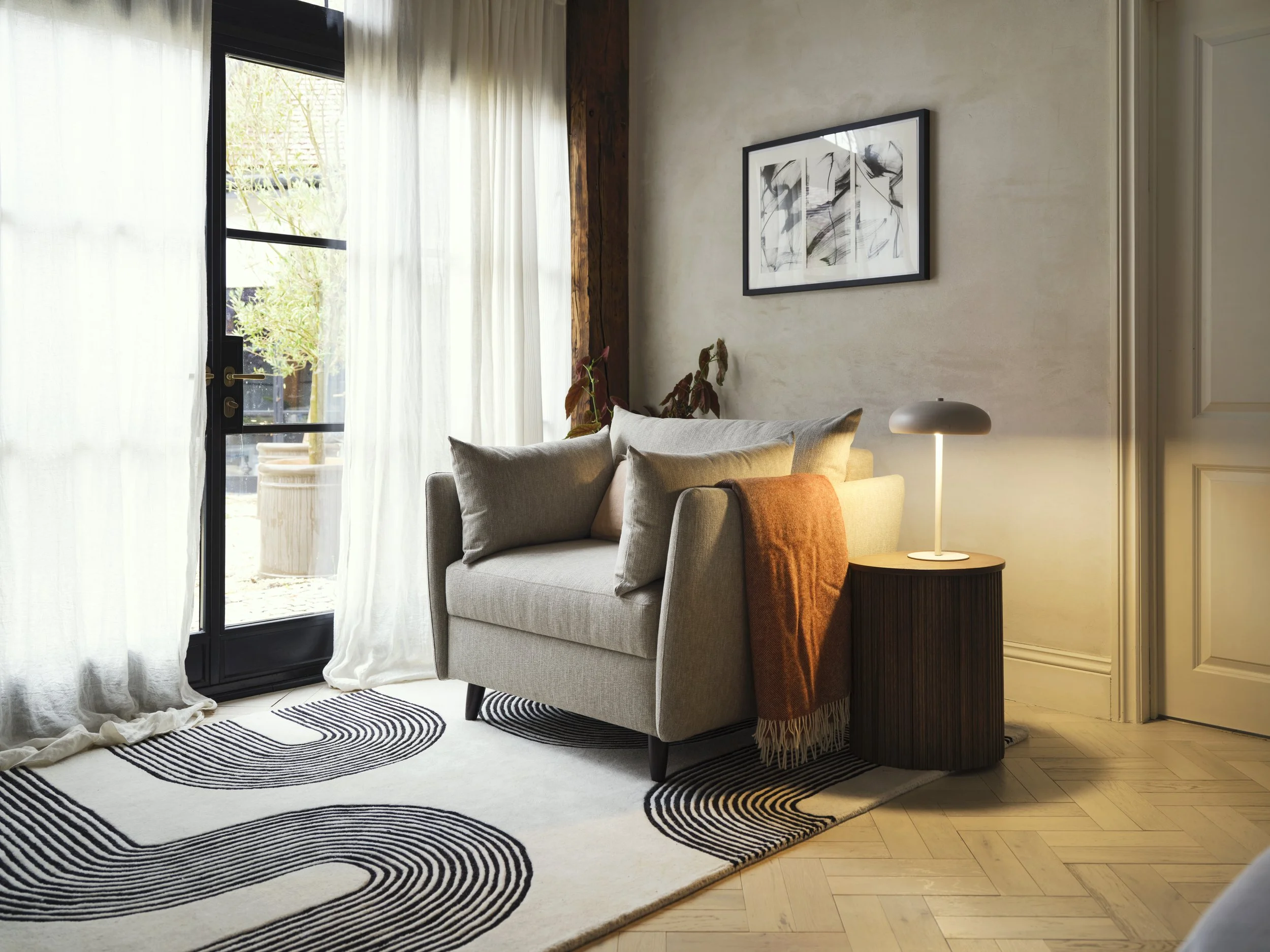 A living room with a beige sofa, a black framed abstract artwork on the wall, a side table with a white lamp, and large windows with sheer white curtains letting in natural light.