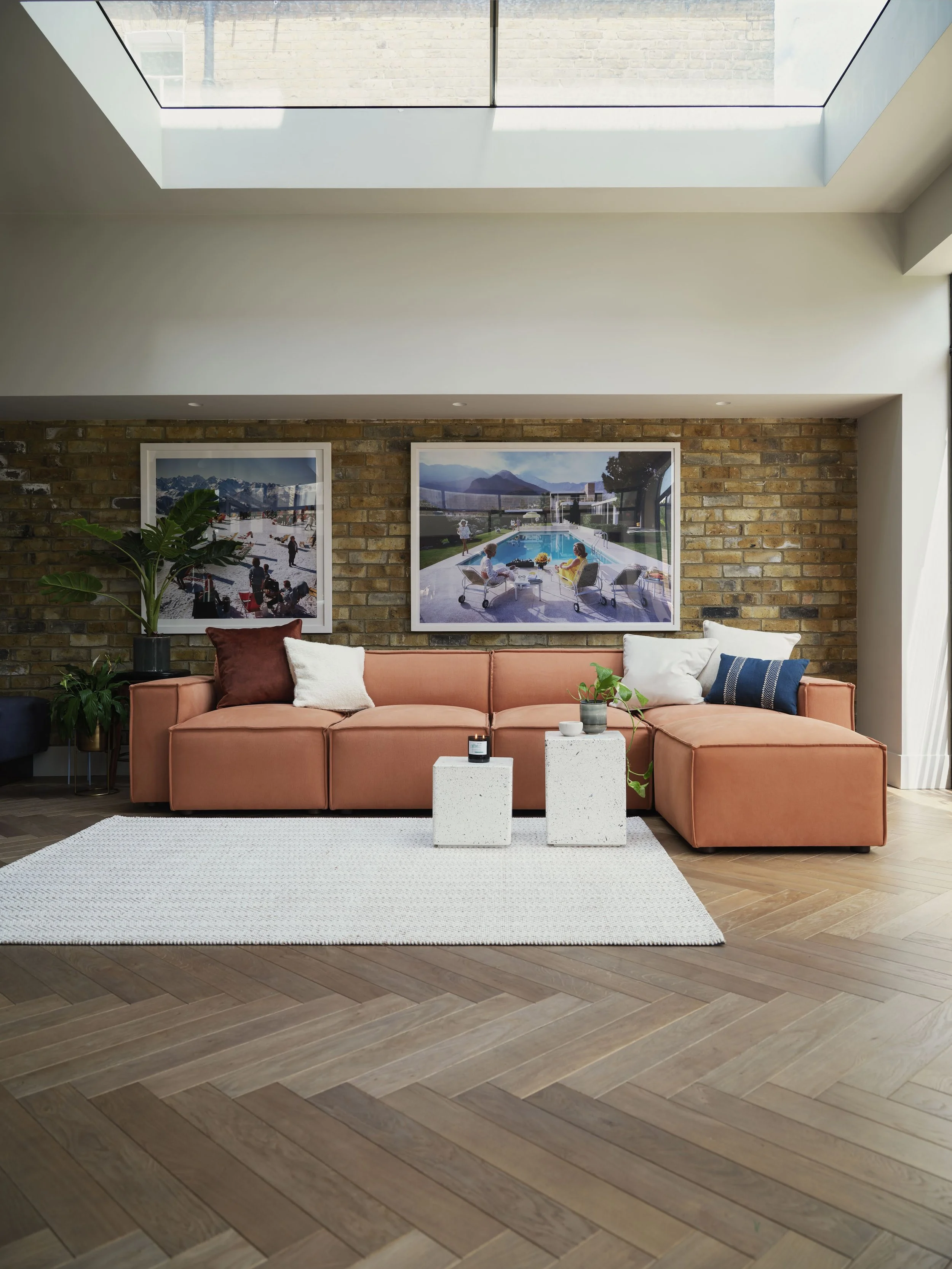 Living room with a sectional sofa decorated with various pillows, next to a white woven rug, a potted plant, and two white cube tables with candles, against a brick wall with two large framed pictures of outdoor scenes. There's a skylight above, providing natural light.