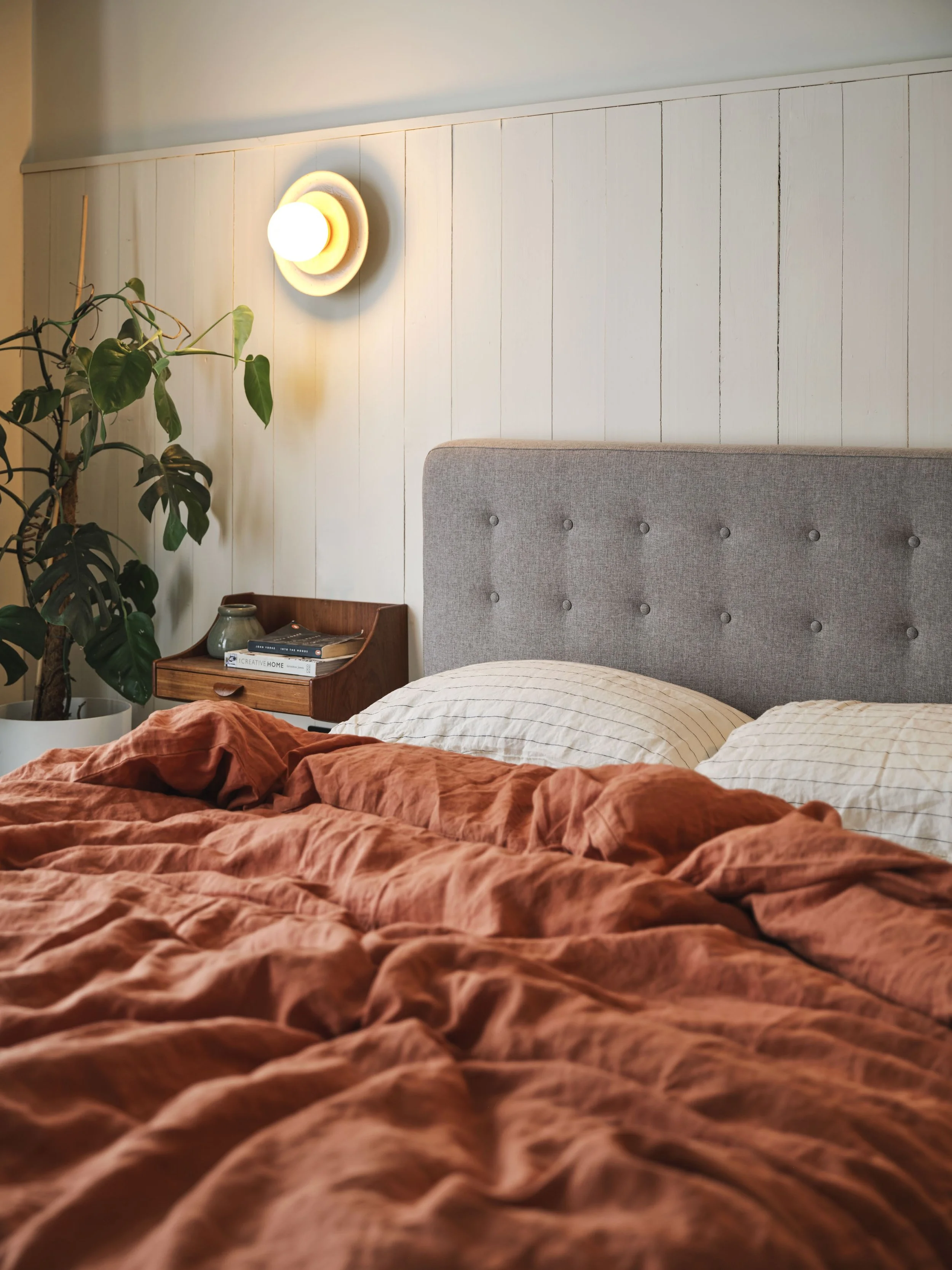 A cozy bedroom with a beige upholstered headboard, a wooden nightstand holding a ceramic vase and books, and a large green houseplant in a white pot. The wall behind features white vertical wooden panels, with a round wall light providing warm lighting.