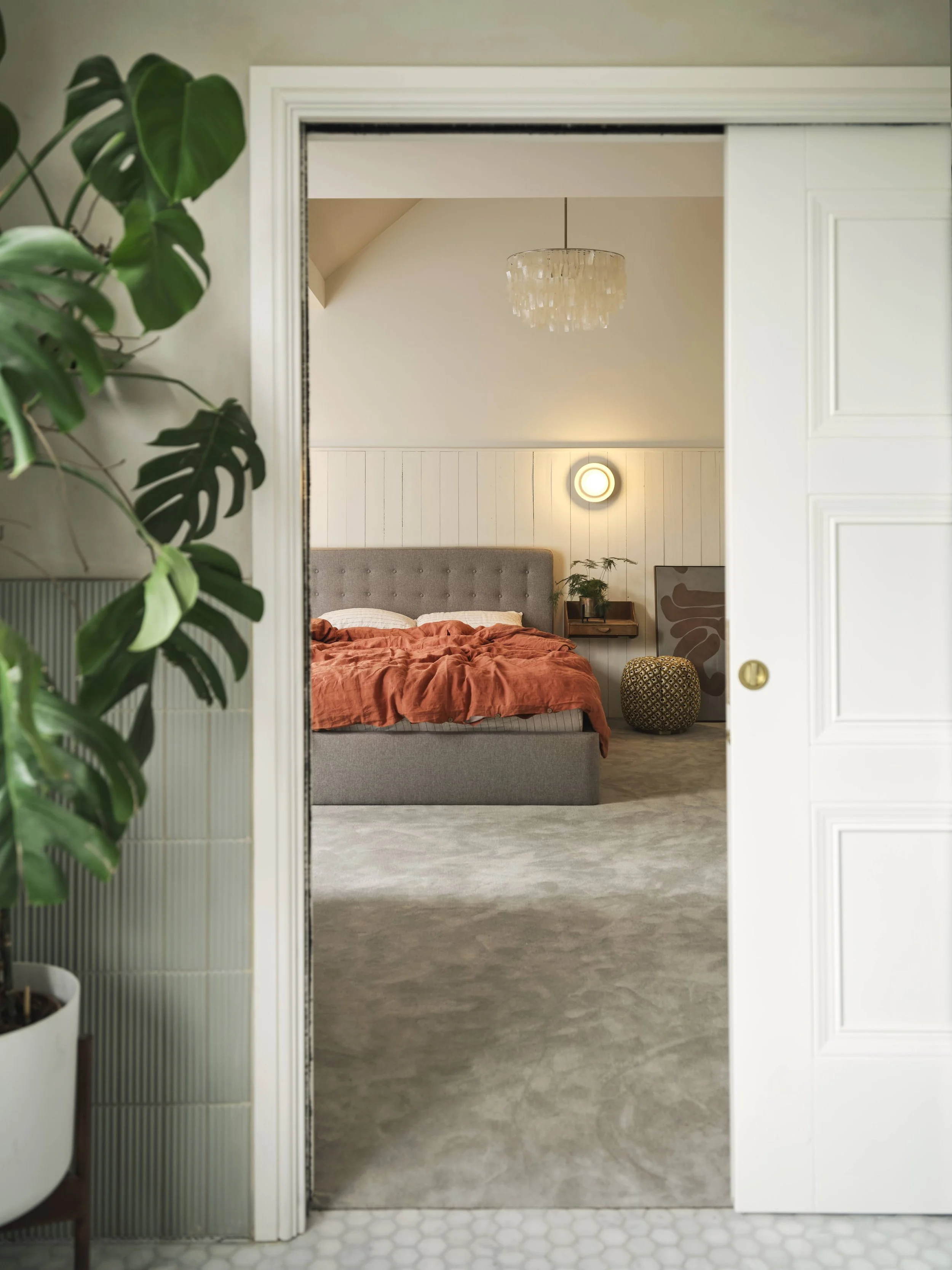View of a bedroom through a partially open white sliding door, with a gray upholstered bed, orange bedding, a small nightstand with plants, a wall-mounted circular light, a chandelier, and artwork. A large green potted plant is visible on the left outside the room.