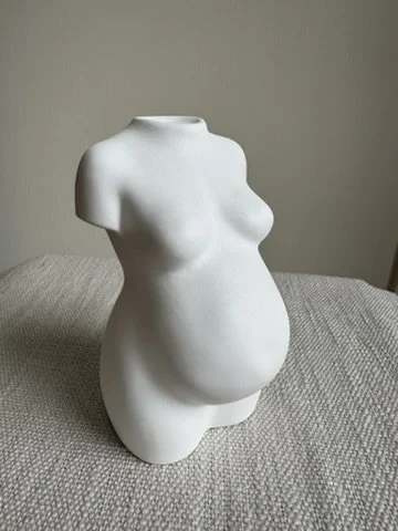 White abstract sculpture resembling a pregnant female torso on a grey surface.