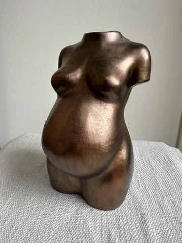 Bronze-colored bust sculpture of a pregnant female torso on a grey fabric surface against a light wall background.