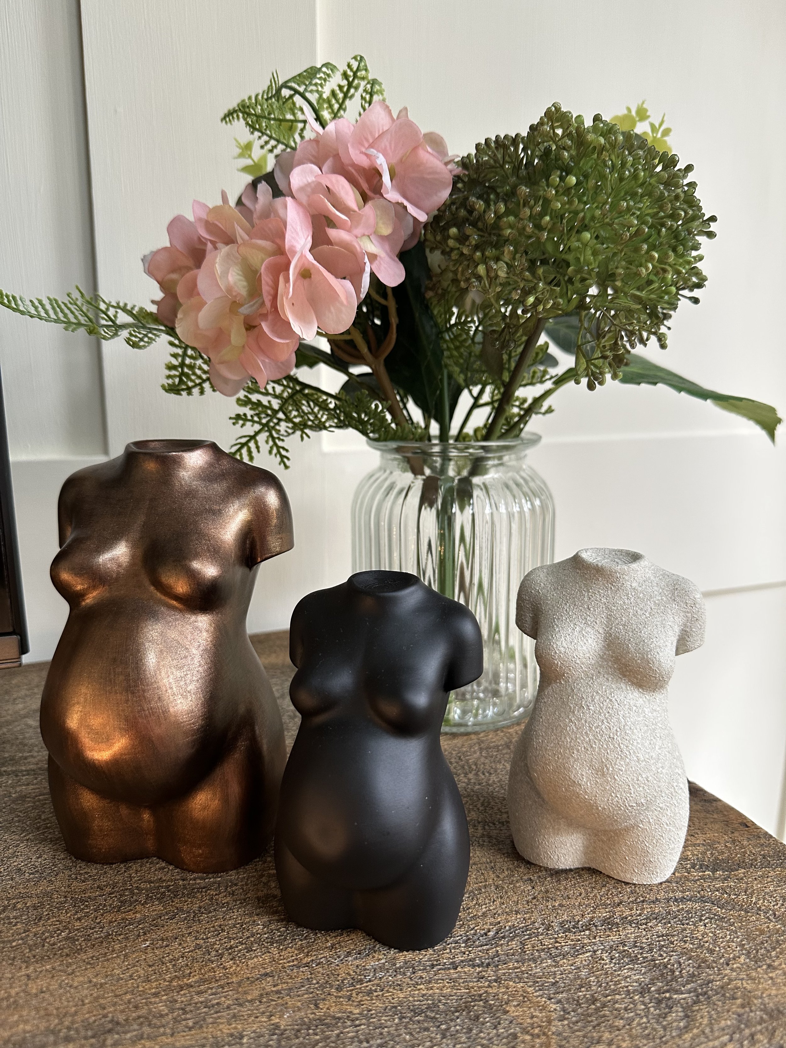 Decorative flower arrangement in a glass vase with pink hydrangeas and green foliage, accompanied by three small sculptures of pregnant women in metallic, black, and textured white finishes displayed on a wooden surface.