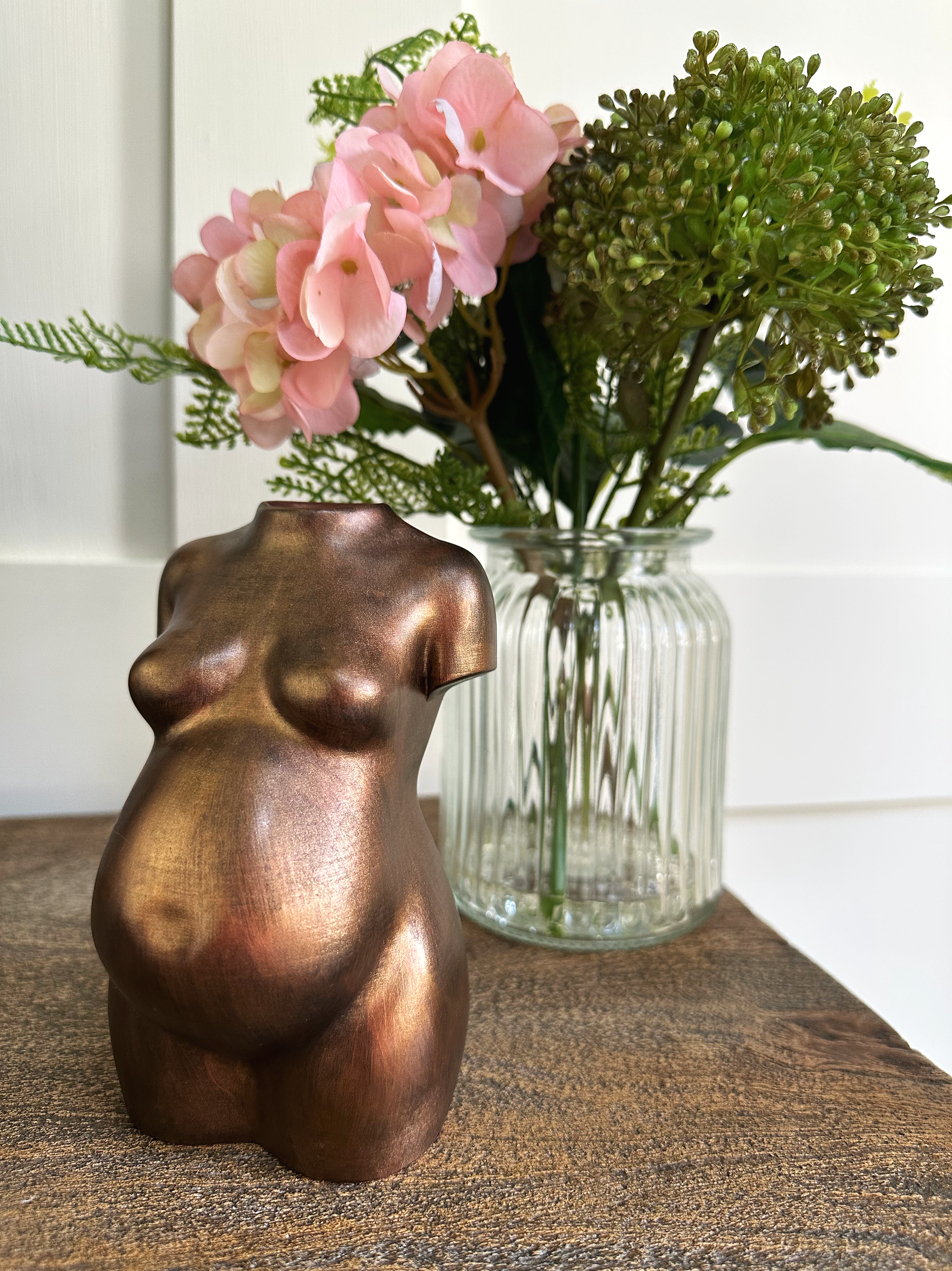 A metallic bronze-coloured sculpture of a pregnant female torso, placed on a wooden surface. Behind it, a clear glass vase holds a bouquet of pink hydrangea flowers and greenery.