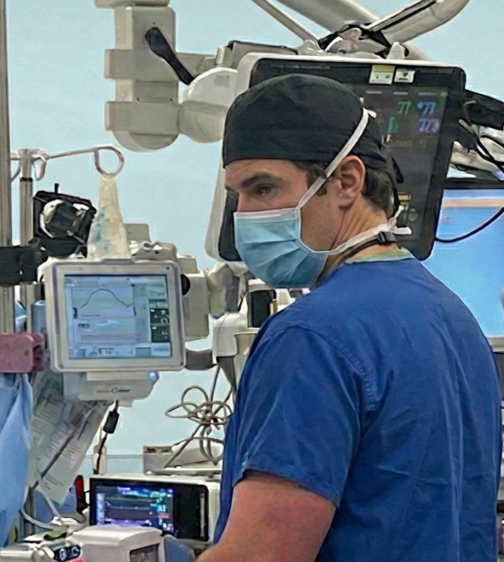 A male surgeon or medical professional in scrubs and surgical cap wearing a face mask, working in a hospital operating room with medical monitors and equipment surrounding him.