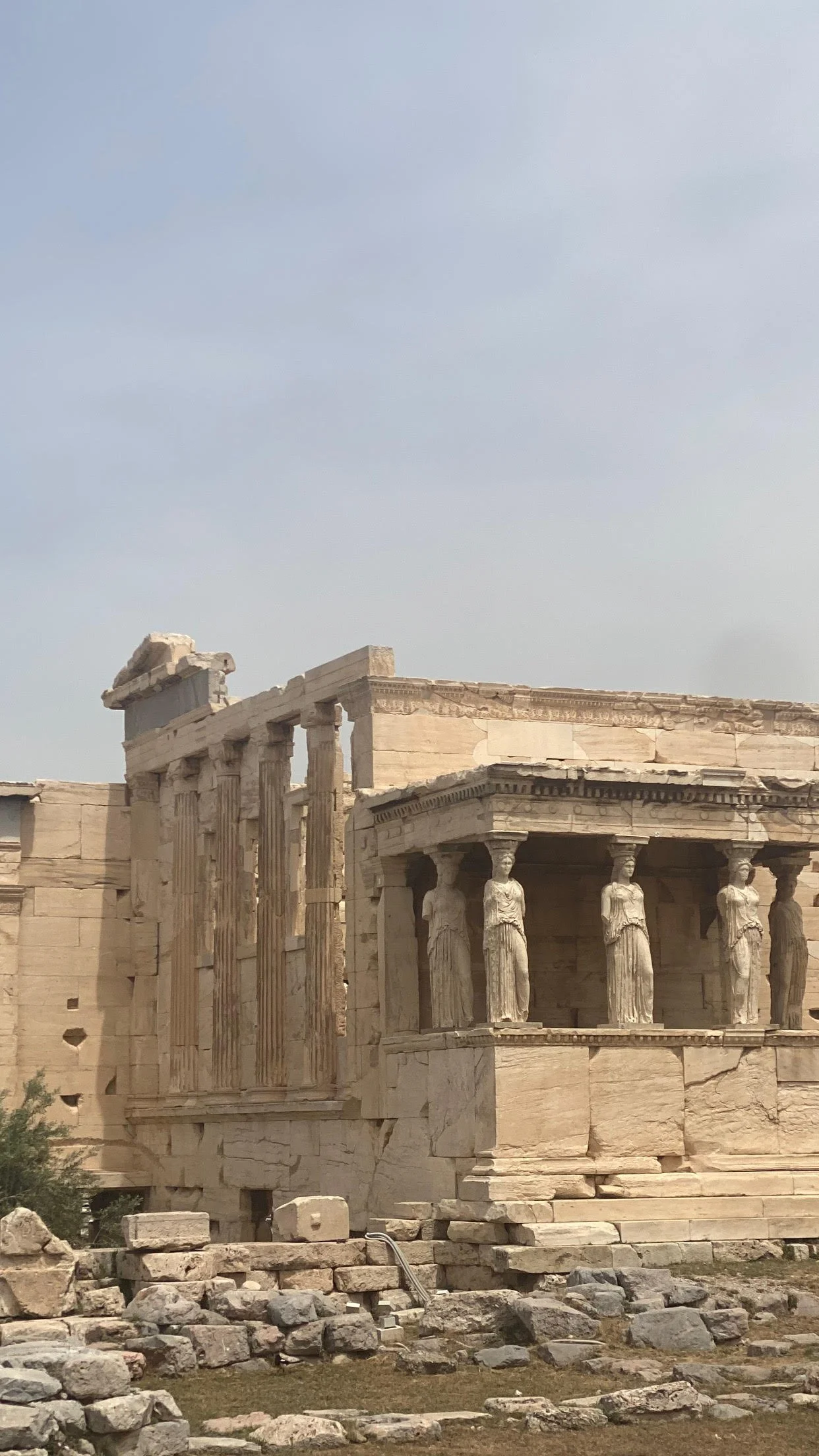 Partial view of the ancient Parthenon temple with damaged columns and statues on a cloudy sky background.